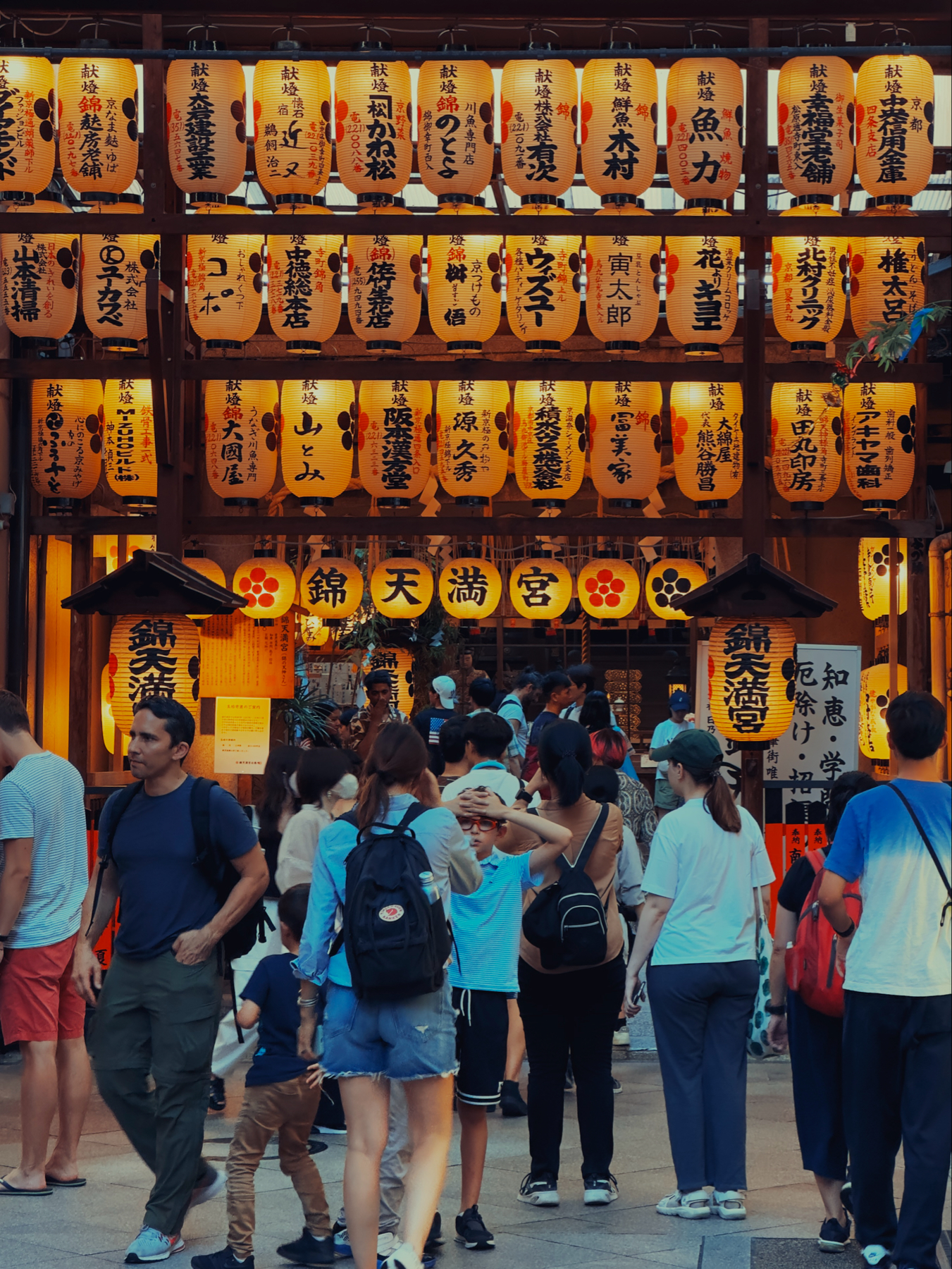 A crowd of people gathered near a Japanese shrine entrance, illuminated by traditional lanterns with Japanese writing.