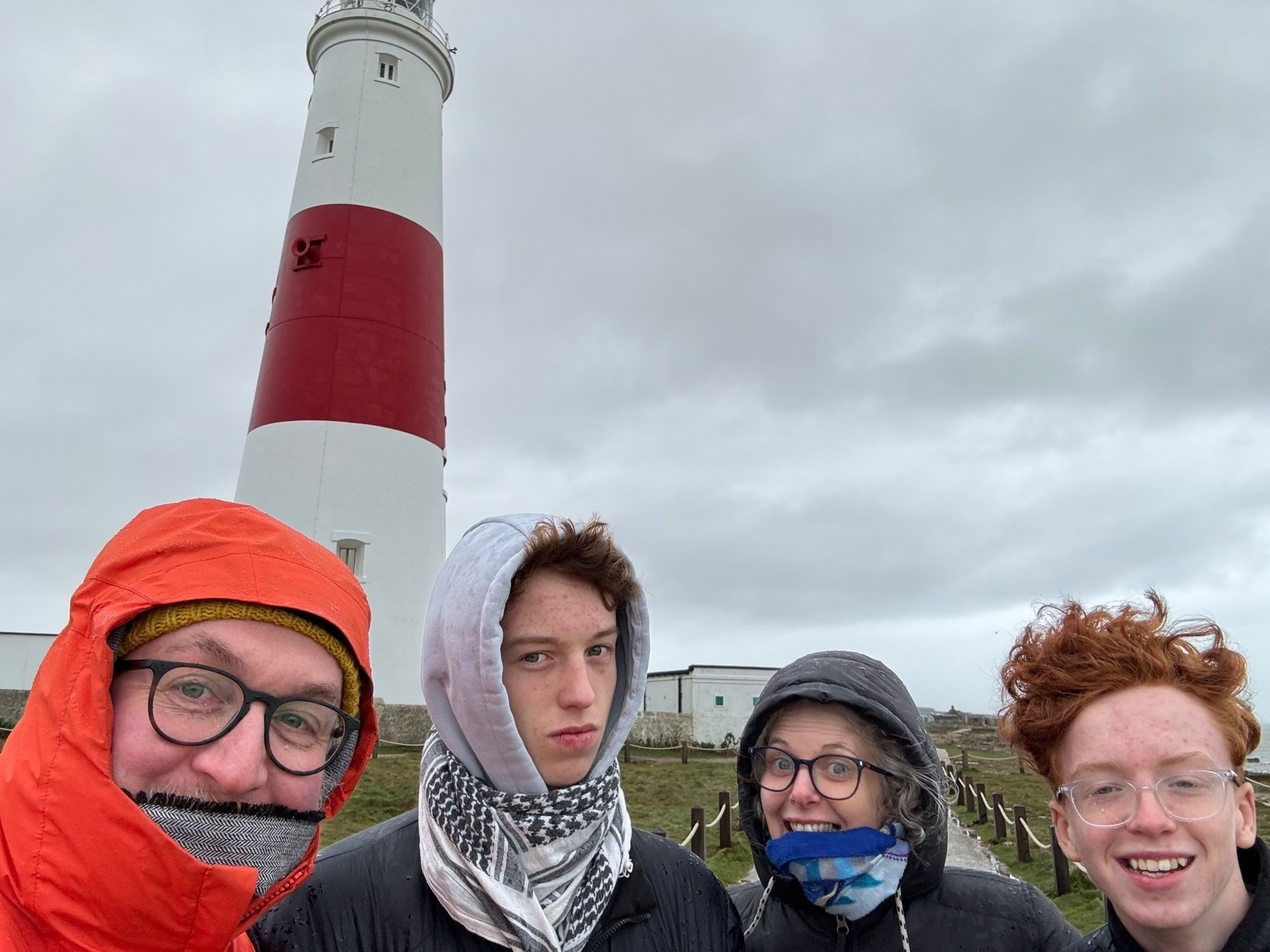 Four individuals standing in front of a lighthouse, with overcast skies in the background. The lighthouse is white with a red band near the top. The people are dressed in cold-weather clothing, with hoods and scarves.