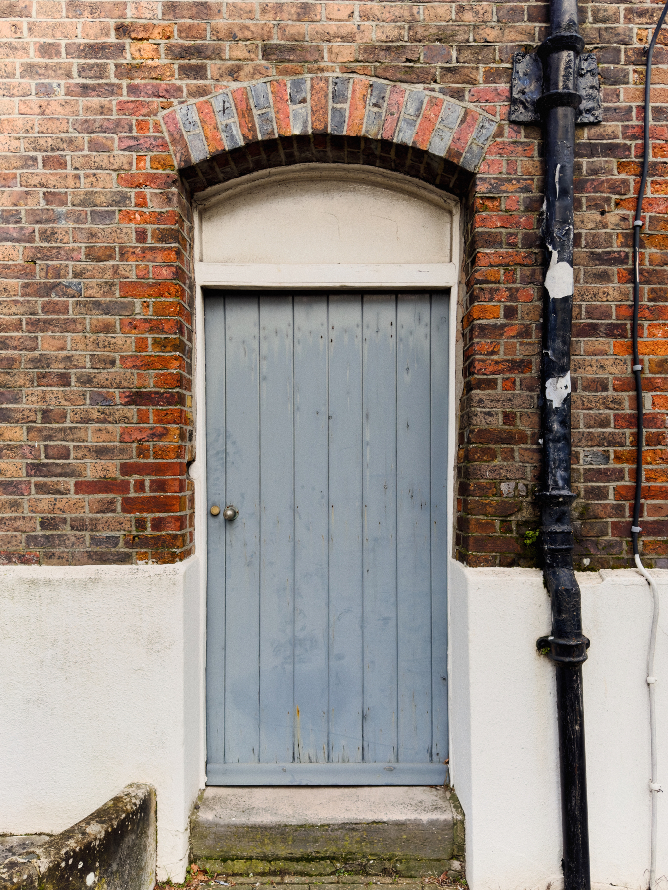 A pale blue wooden door set within a brick wall, with an arched brick lintel above and a cast iron downpipe to the right.