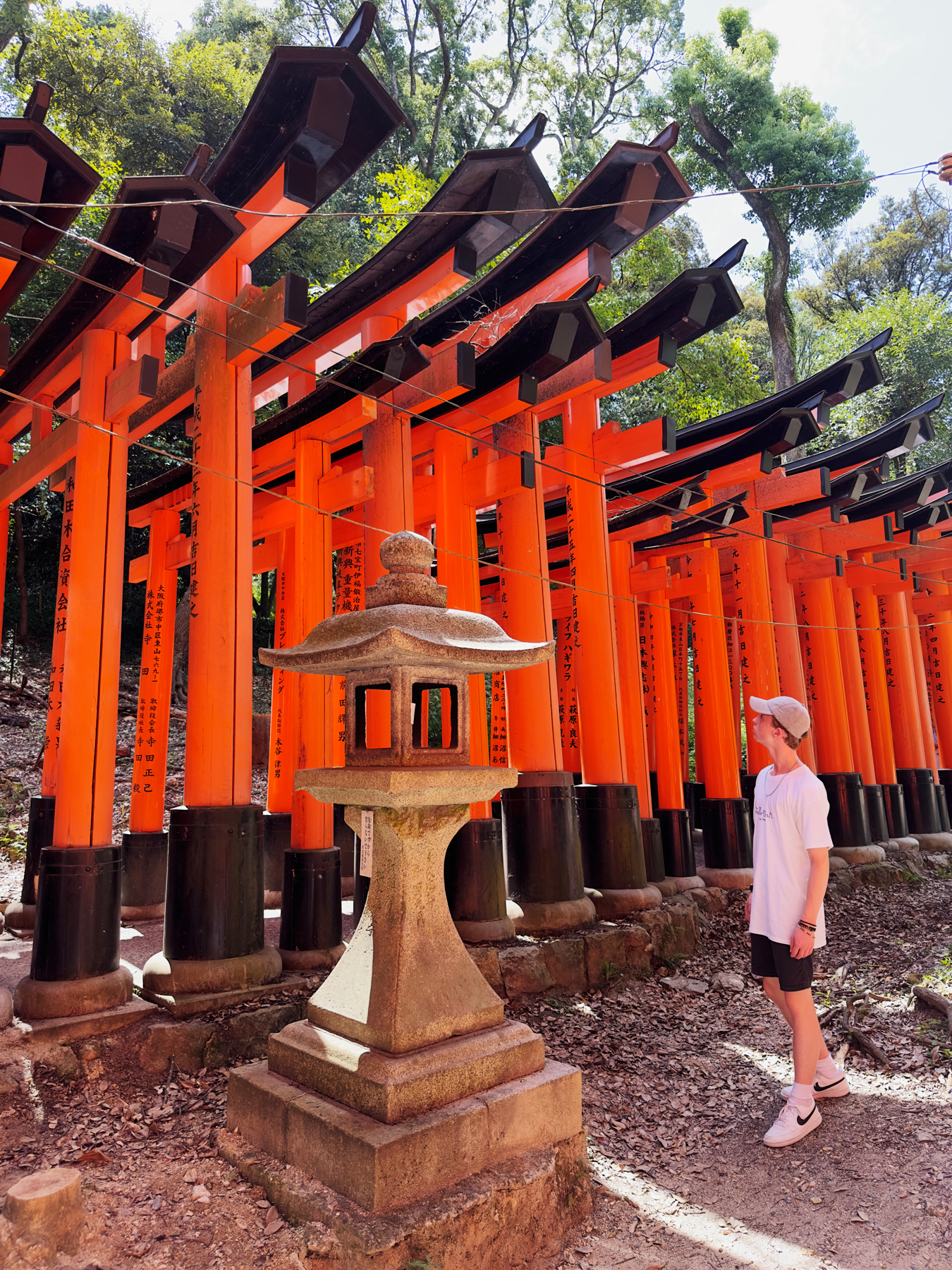 My eldest Son standing next to a traditional stone lantern, with a background of vibrant orange torii gates at Fushimi Inari Shrine in Kyoto, Japan.