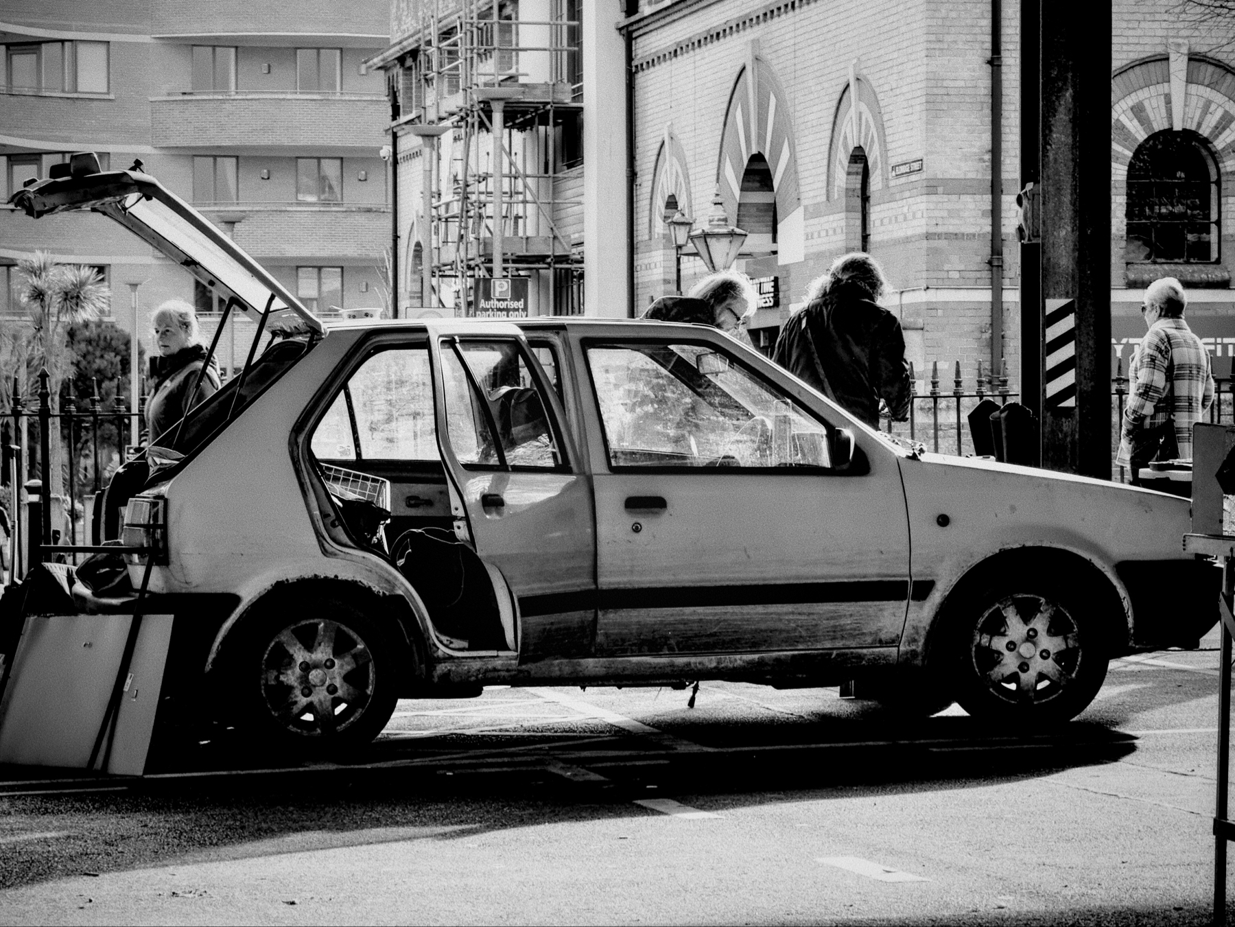 A black and white photograph of a heavily damaged car parked on a street with broken and missing parts, including doors and windows, with people walking by in the background.