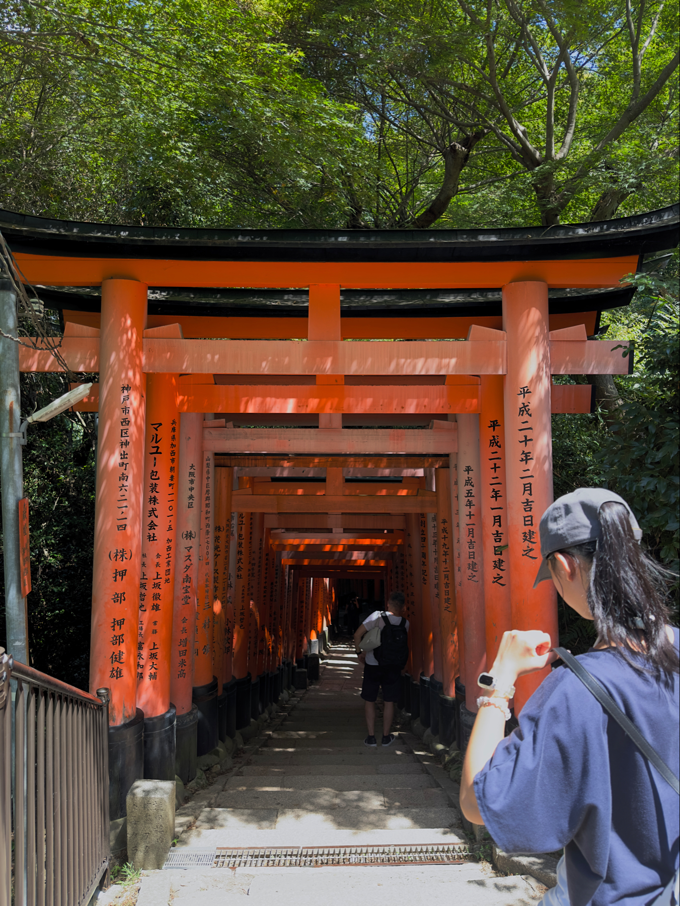 A tree-lined pathway leading through a series of traditional Japanese torii gates at Fushimi Inari Shrine in Kyoto, with visitors walking beneath the vermilion structures.
