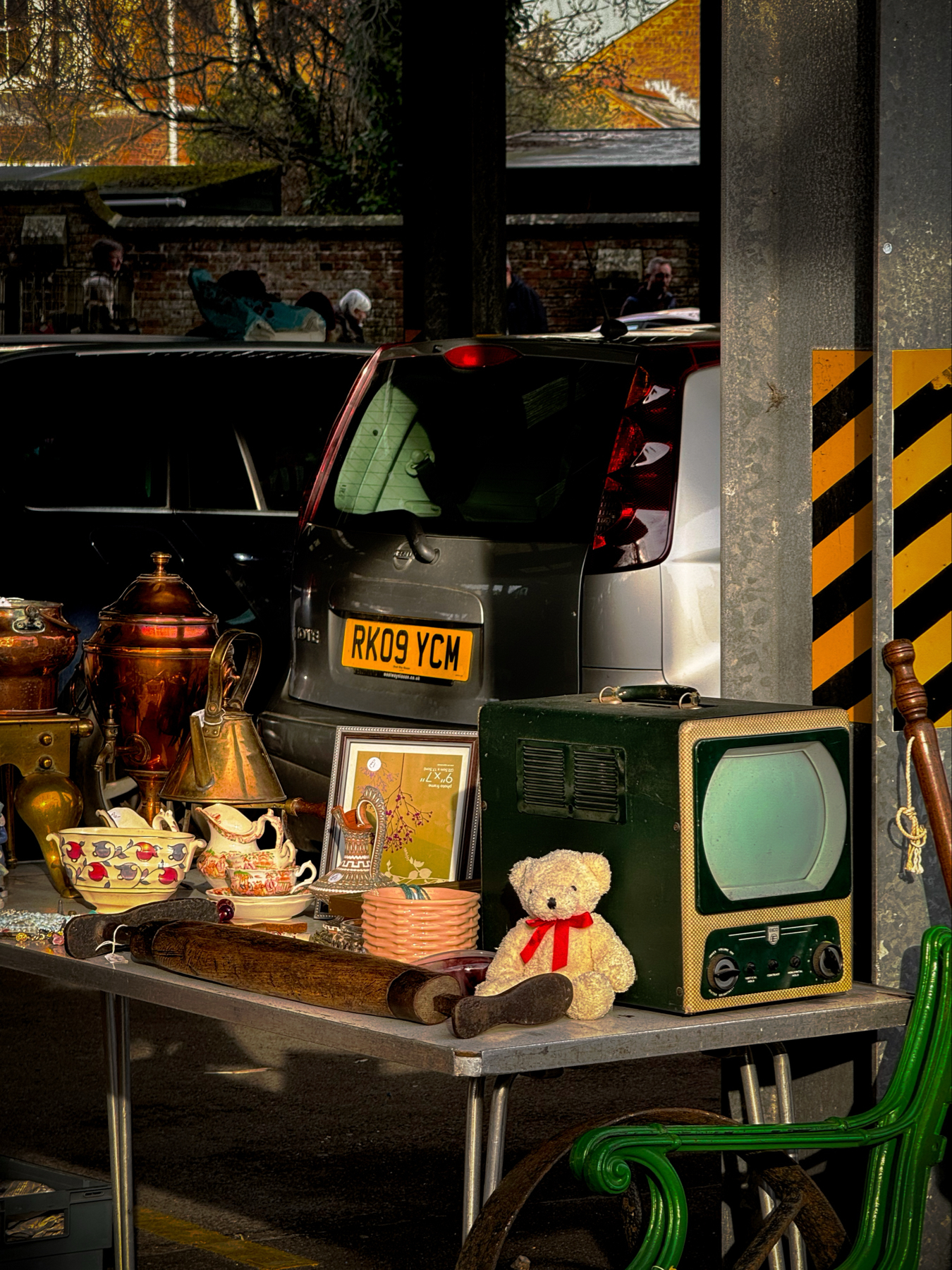 A variety of vintage items displayed on a table at a flea market, including a teddy bear, an old television, assorted pottery, and metal teapots, with parked cars and people in the background.