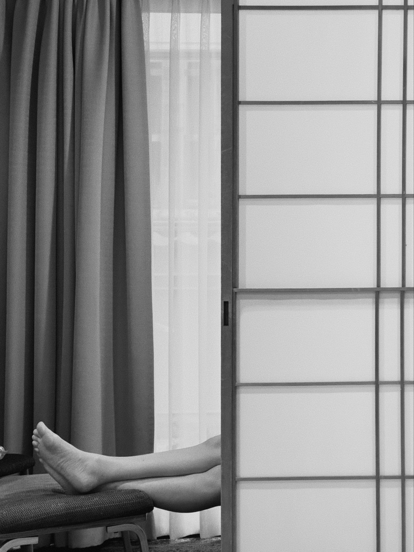 Black and white photo showing the lower legs and feet of a person reclined and resting on a chair, partially obscured by a curtain, next to a door with frosted glass panels.