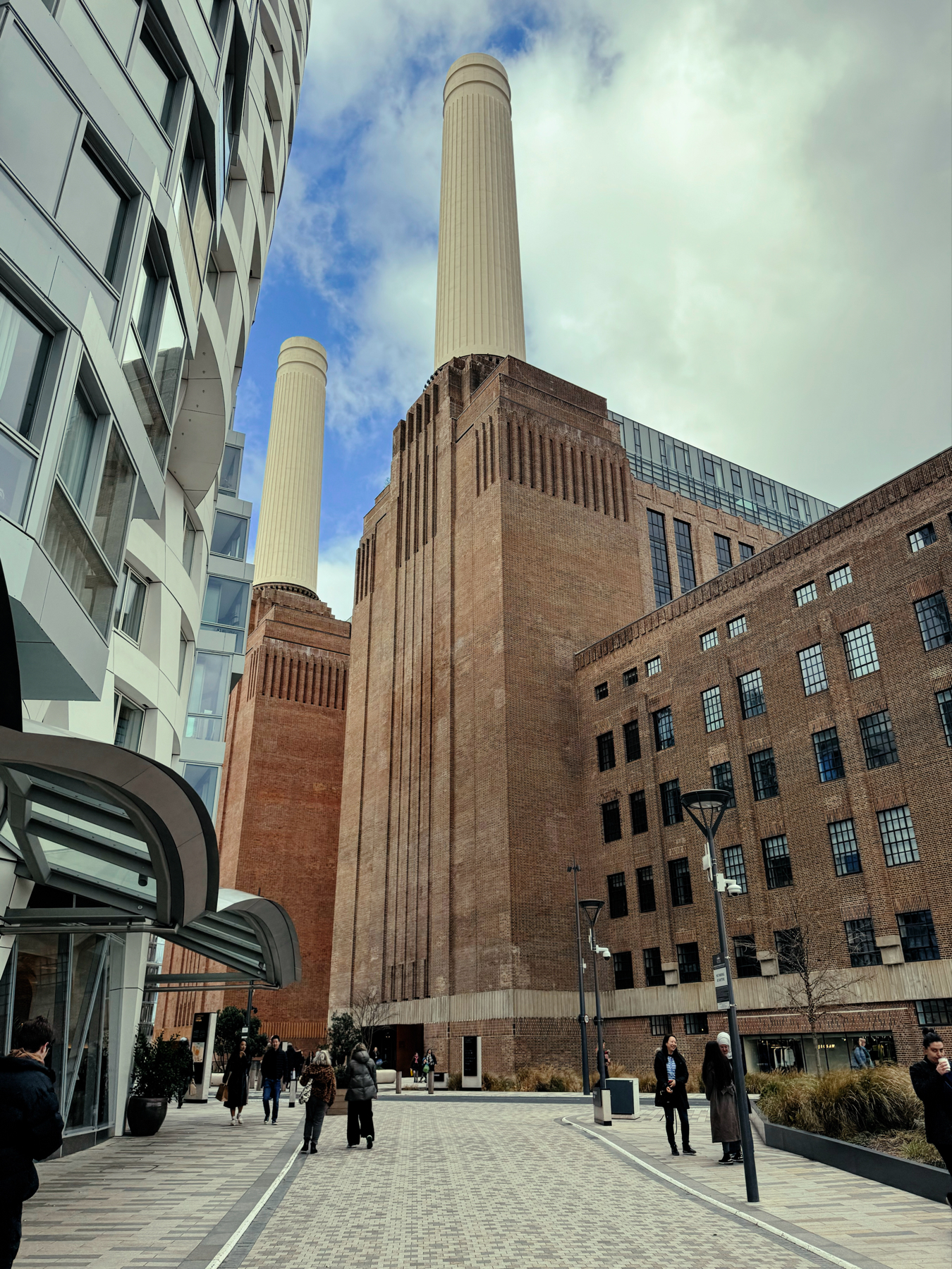 A cityscape featuring the iconic brick facade and tall chimneys of Battersea Power Station with people walking on the paved area in front and contemporary buildings to the side. Clouds adorn the sky above.