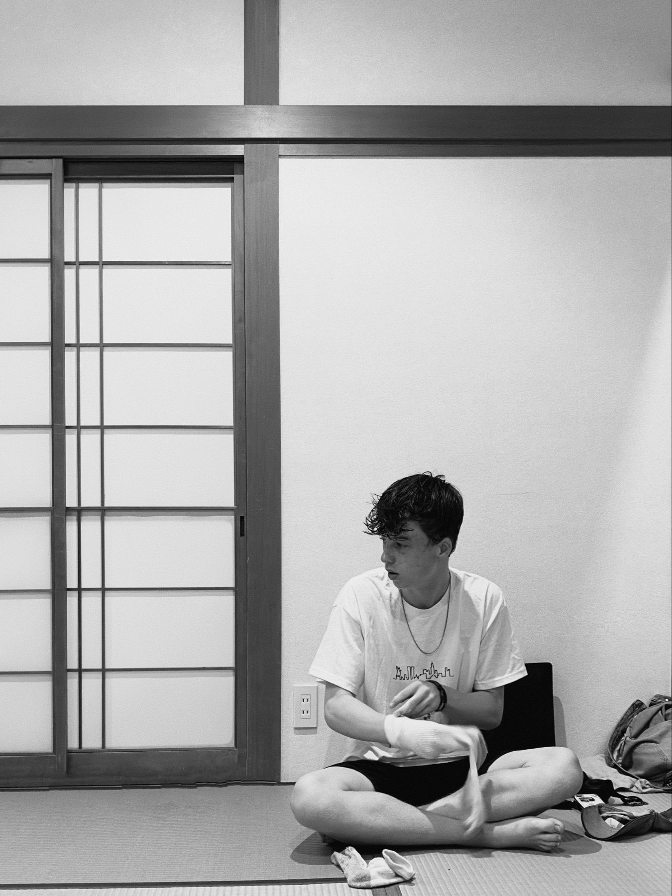 A person sits cross-legged on a tatami floor in a traditional Japanese room with shoji sliding doors. The individual appears pensive and is dressed in casual, modern clothing, contrasting with the room&rsquo;s cultural aesthetic. Black and white photo.