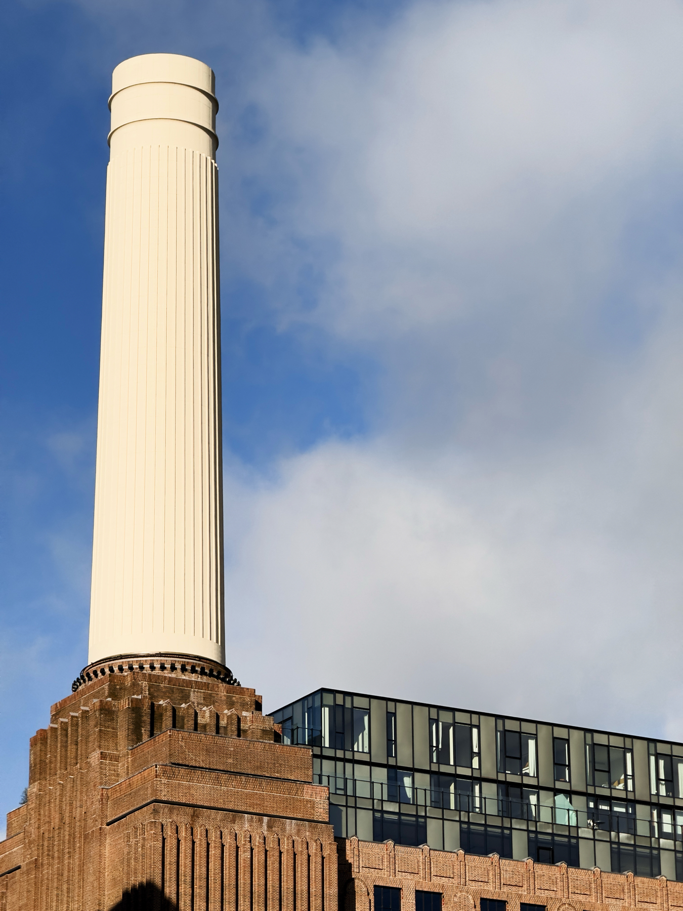 A large, cylindrical white chimney stack rises above a brick building with modern glass extensions, against a blue sky with light clouds.