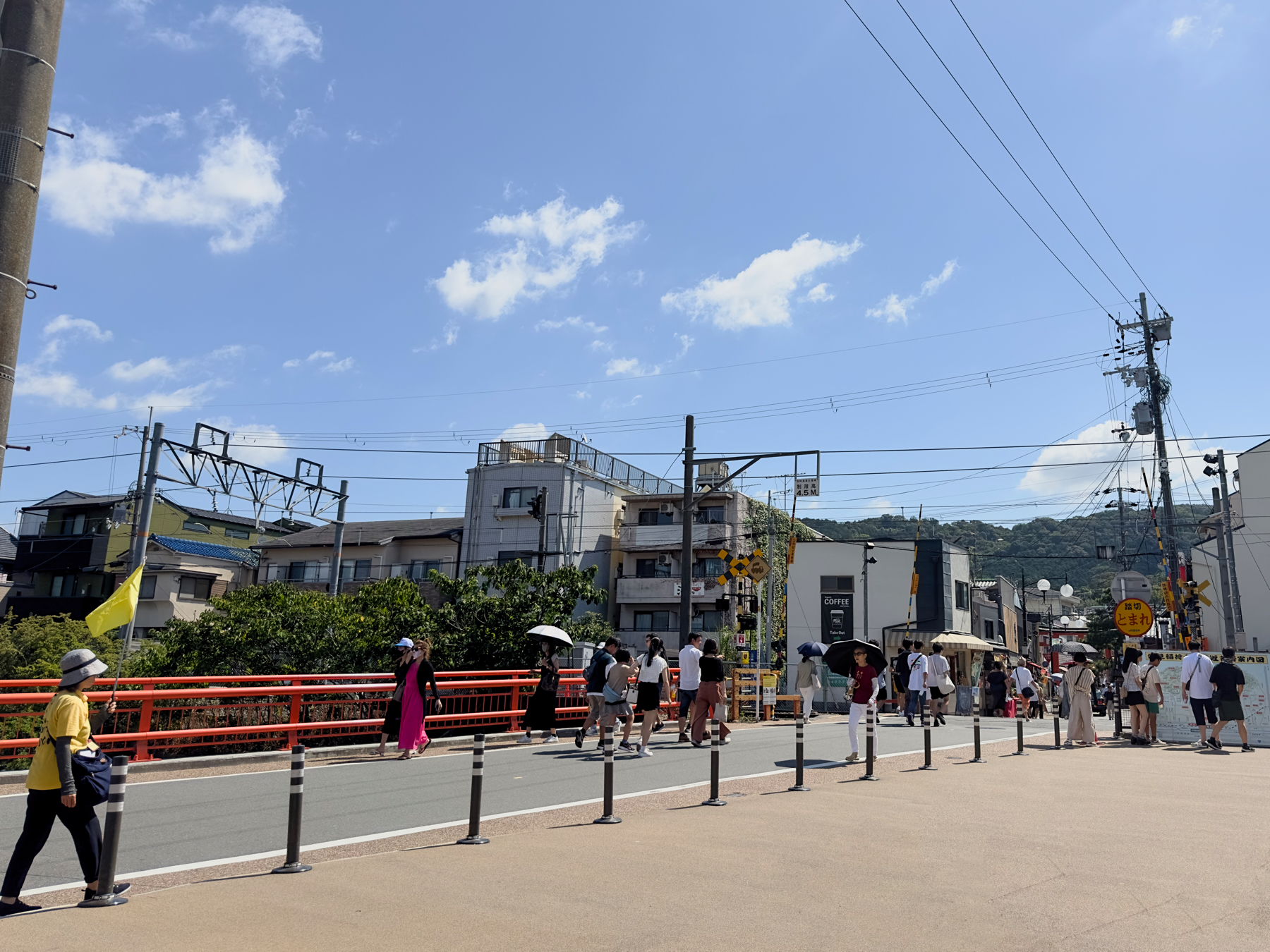 A bustling street scene with pedestrians, some holding umbrellas for shade, walking along a sidewalk with a red railing. There are buildings in the backdrop under a blue sky with scattered clouds. Utility poles and power lines are also visible.