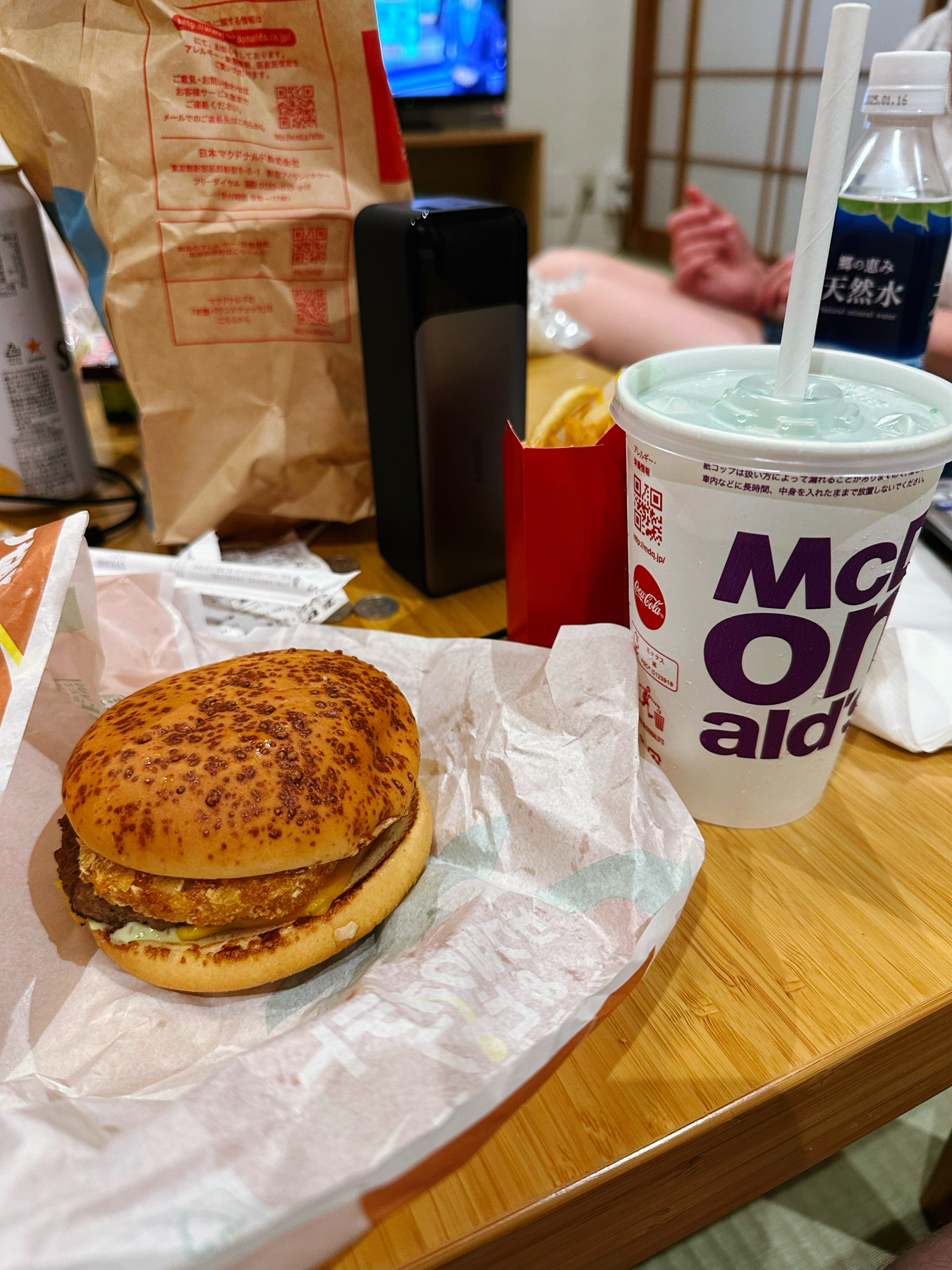 A fast food meal on a wooden table, including a burger, a drink with a straw, French fries, and various takeout packaging, with a blurred background of a living room.