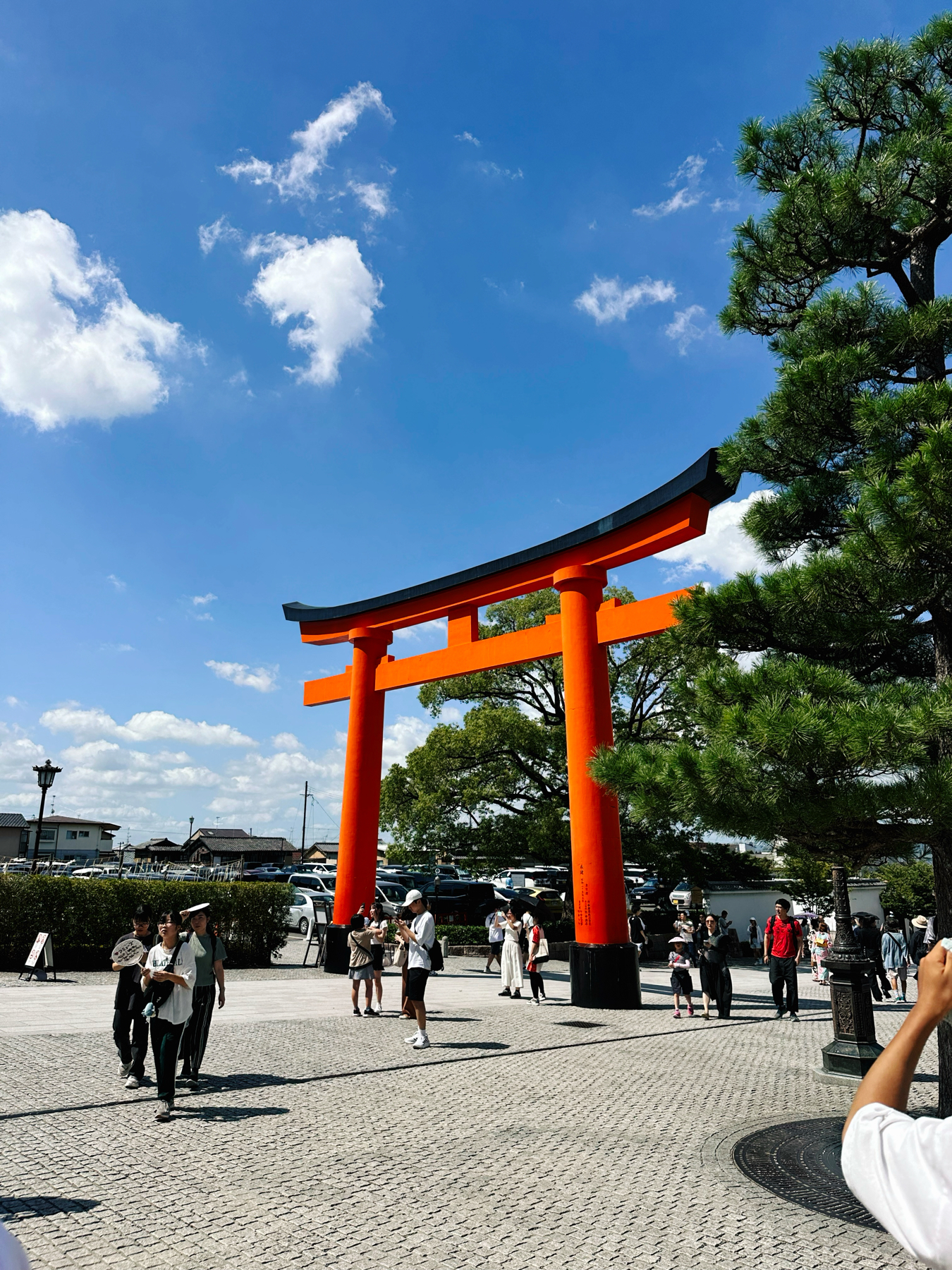 Torii gate at a Japanese shrine with visitors walking around on a sunny day, with clear blue sky and fluffy clouds overhead.