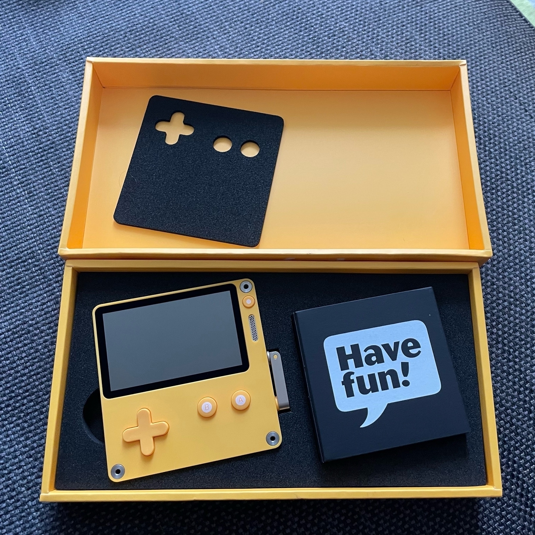 An opened yellow cardboard box, revealing the Playdate game console and a small box of accessories that say "Have fun!" on the front.