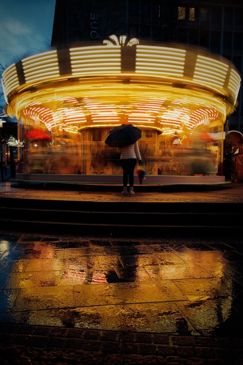 A carousel moving with motion blur. A reflection of the carousel visible on the wet stones in front of it. A person with an umbrella standing before the carousel.