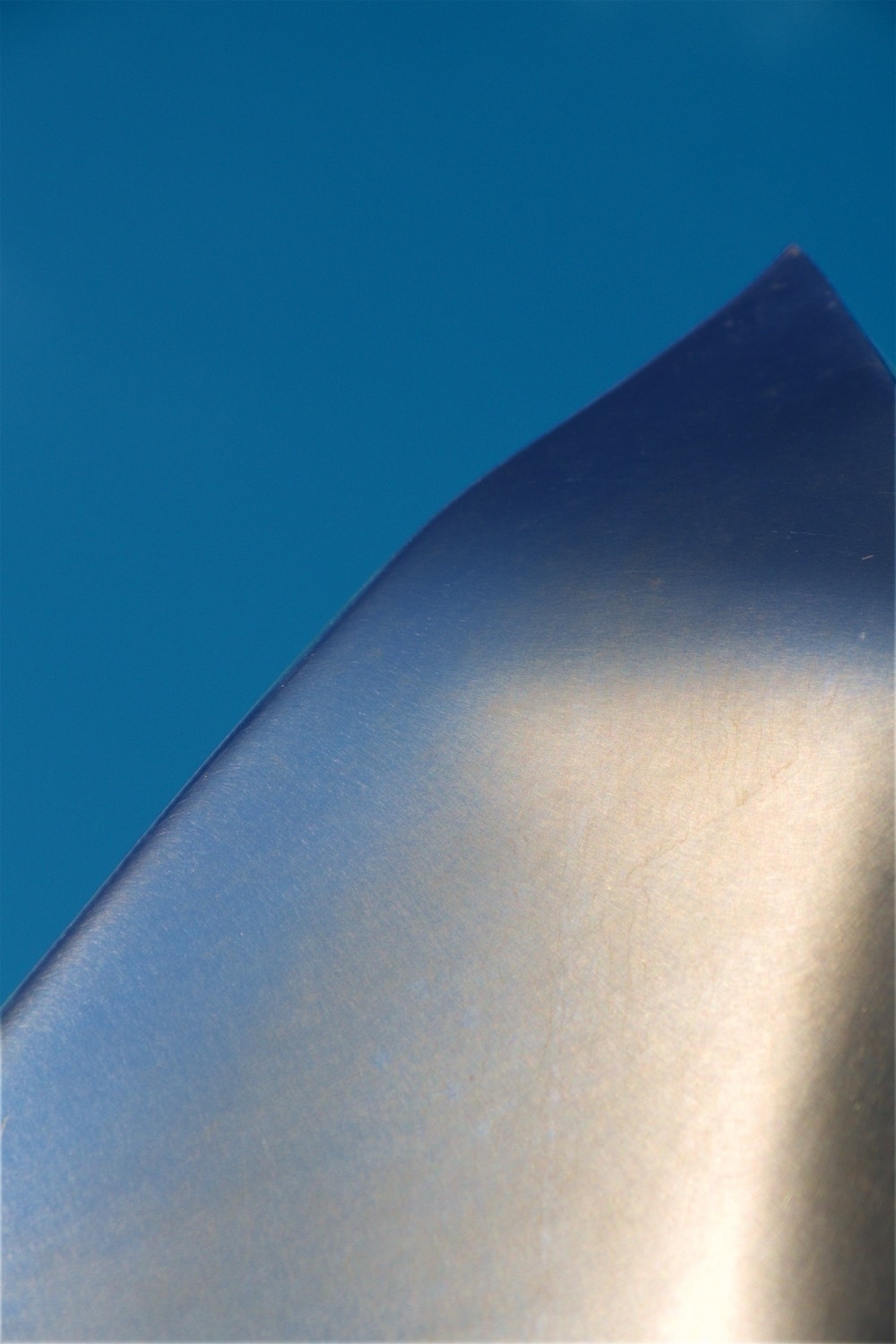 A close up of a steel sculpture. The background is a uniform, blue sky. The sun is reflecting in the sculpture in yellow and orange. Scratches in the steel give some structure to the image.