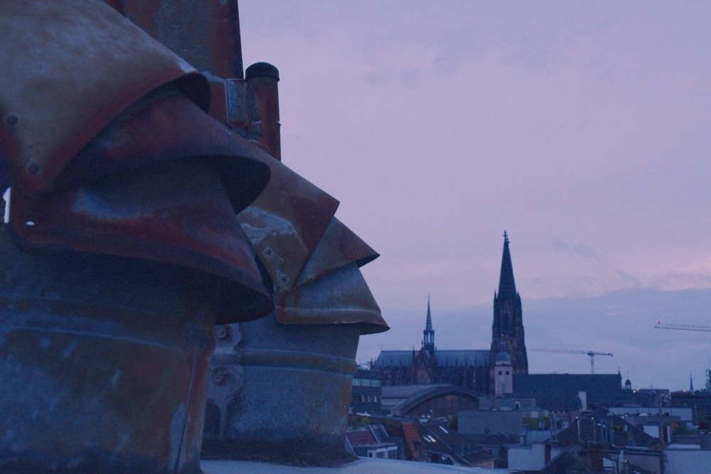 Close-up of a chimney on a roof during the blue hour. In the background rooftops and the Cologne Cathedral.