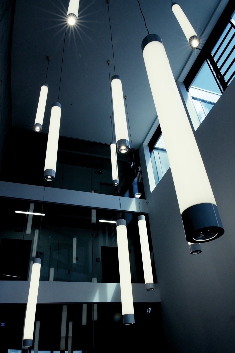 Several rod lamps hang from the ceiling at different heights in a multi-storey room. The lamps also have a spotlight facing down, from which the light radiates in the shape of a star.