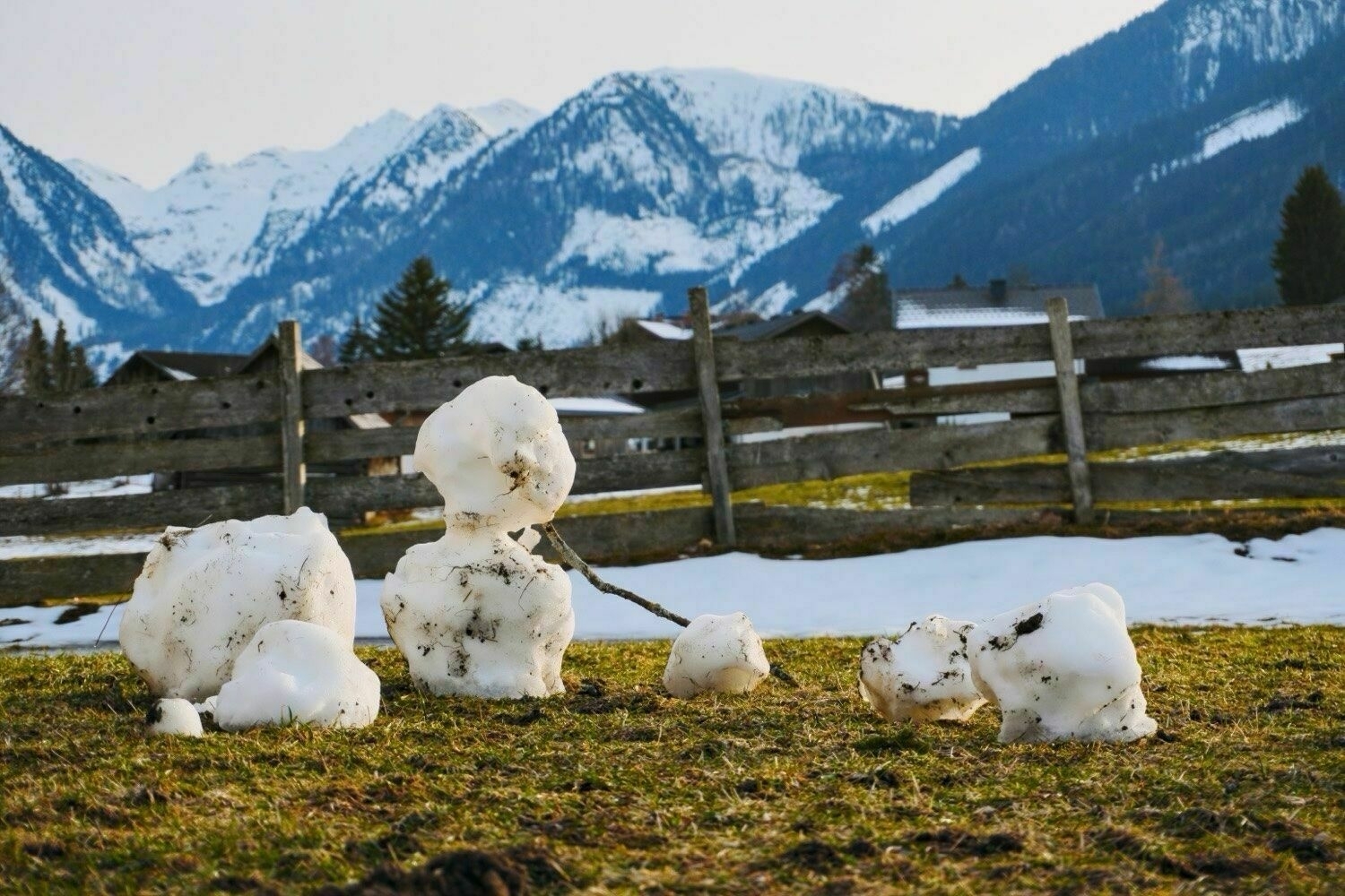 Small partly melted snowman on a green fields with some sopots of snow. Mountains are visible in the background.