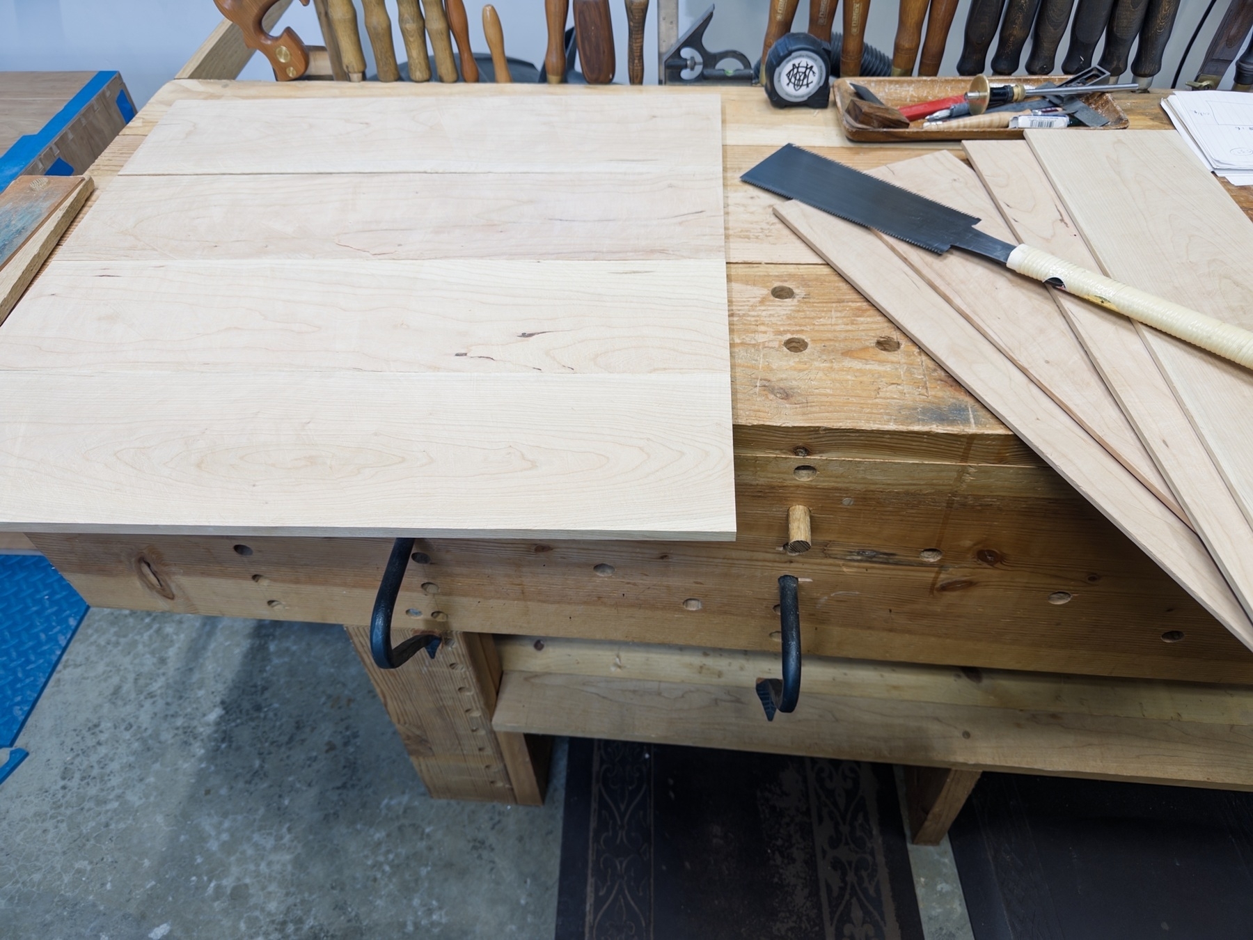 A woodworking bench holds various hand tools, a large wooden board, and several wood panels in a workshop setting.