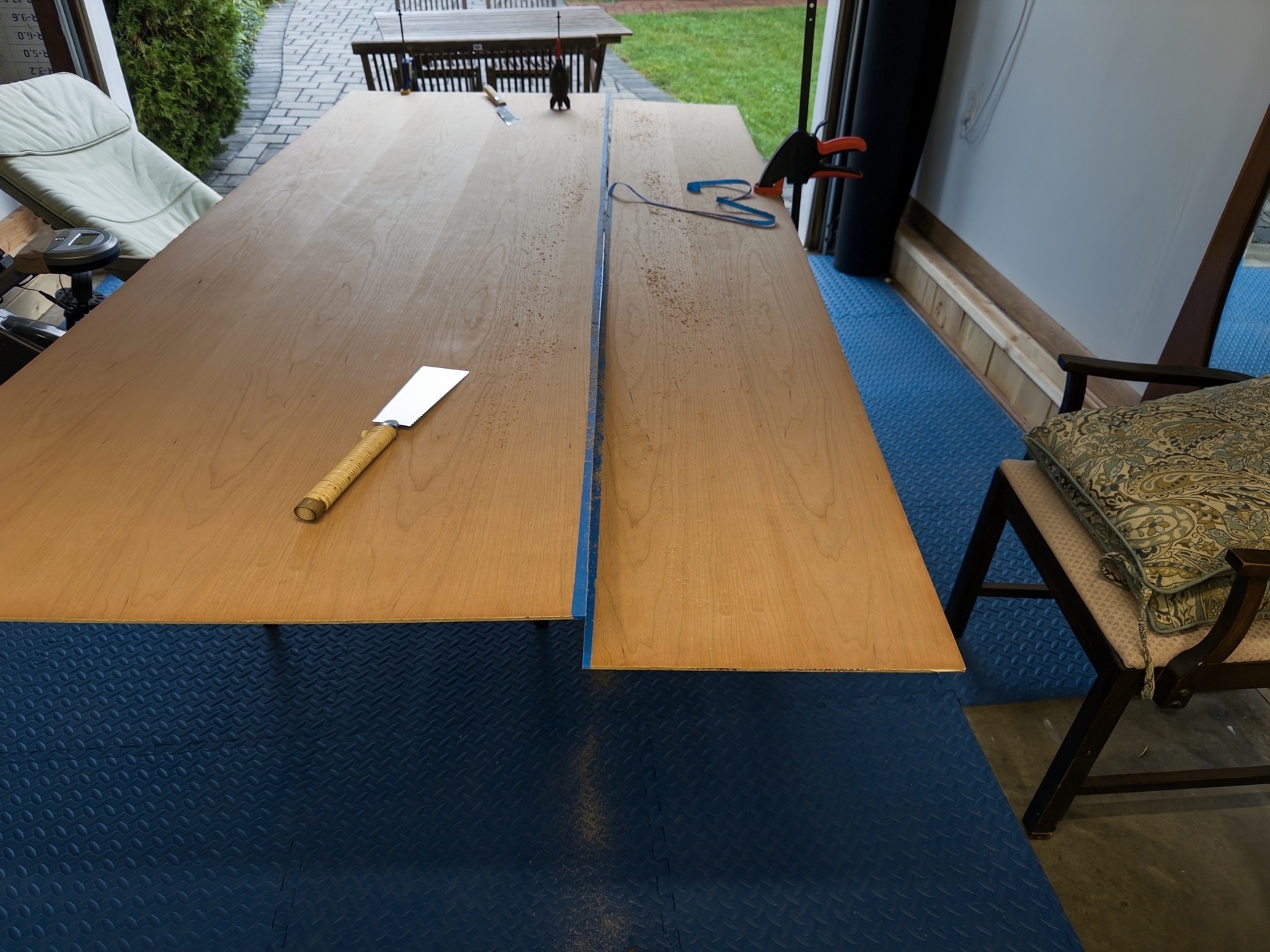 A plywood sheet on table with woodworking tools on a blue floor surface, adjacent to an open doorway leading outside.