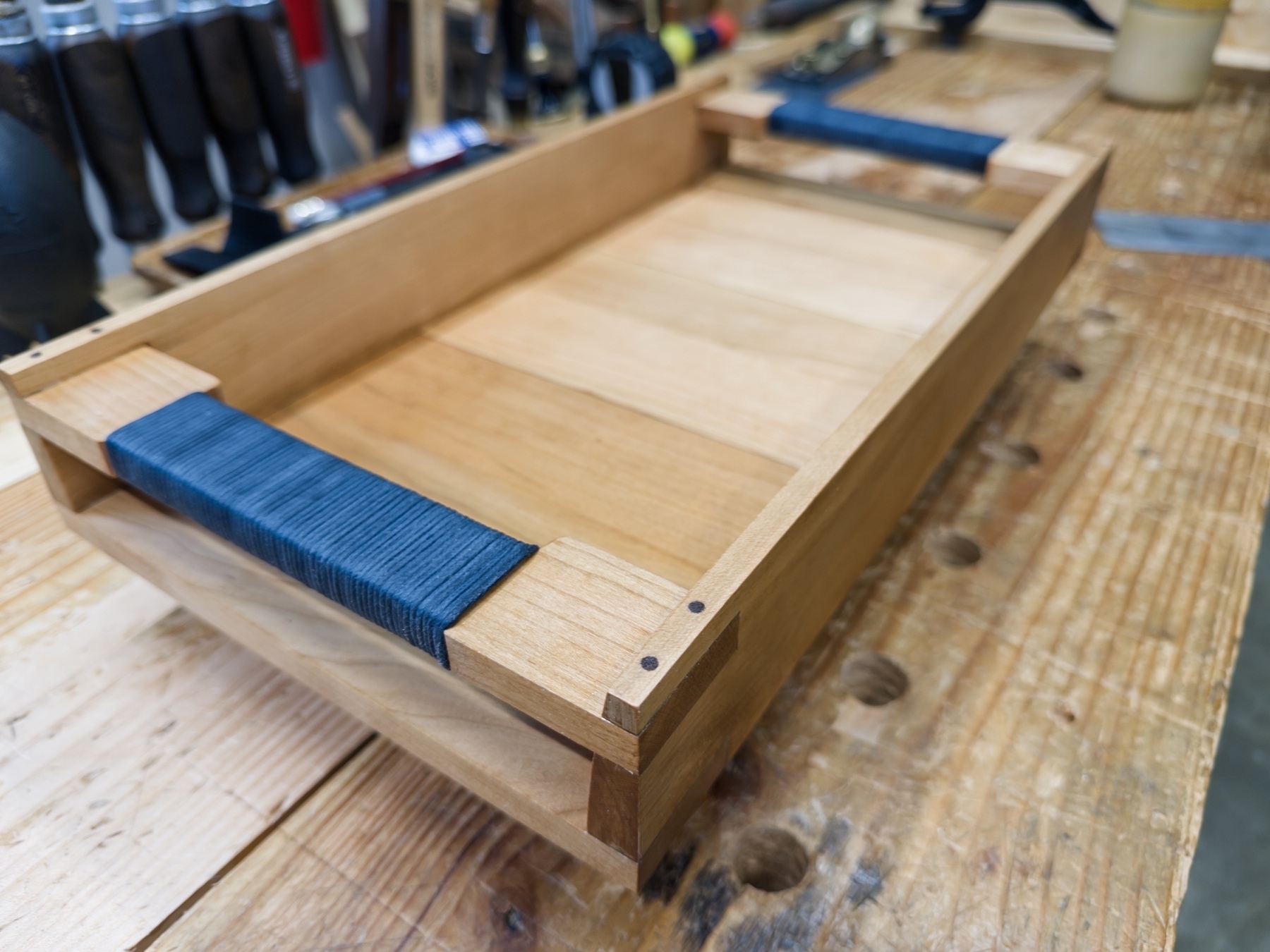 A wooden tray with dark blue handles sits on a workbench surrounded by tools.