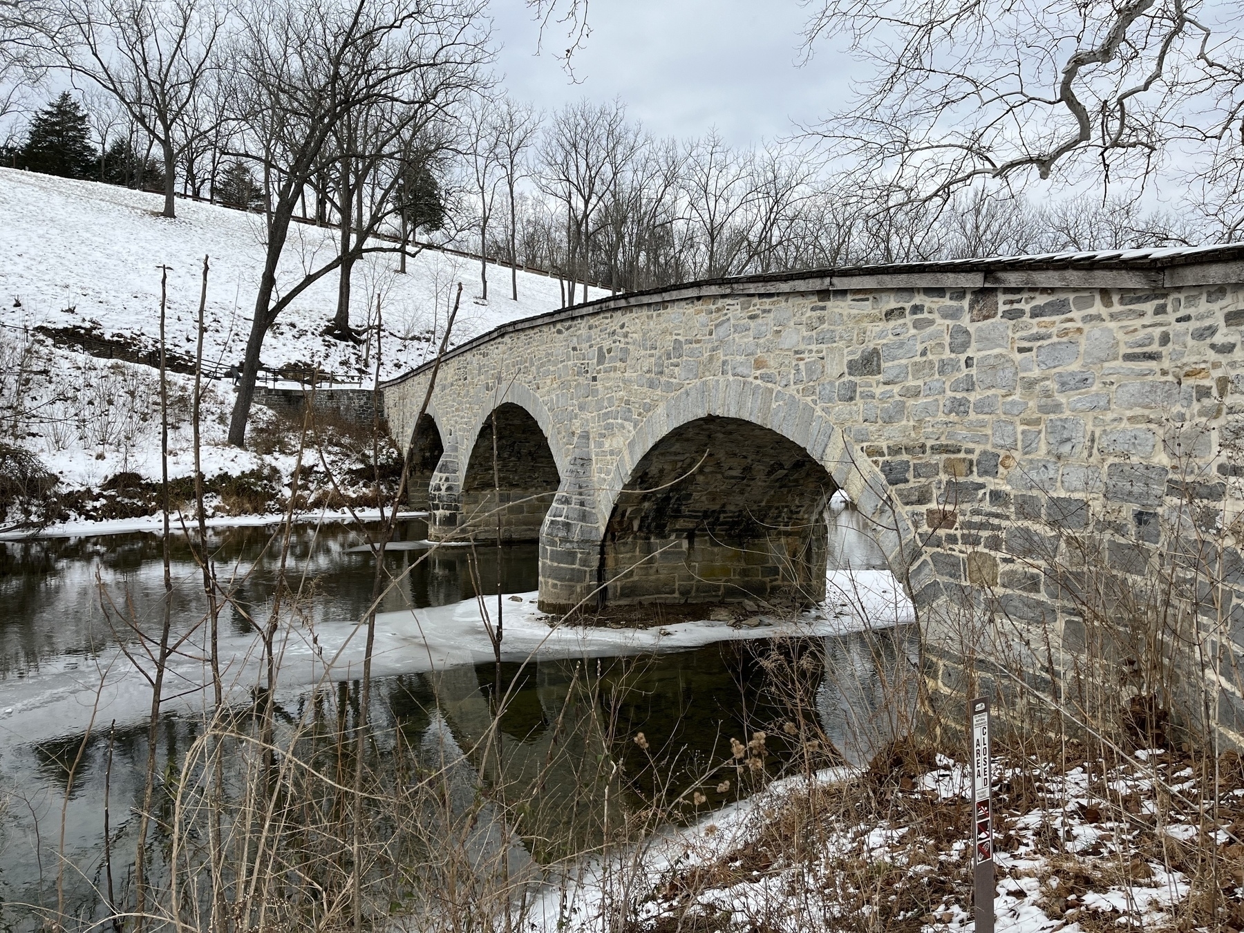 A stone bridge with arches spans a partially frozen river, surrounded by snow-covered trees and landscape.