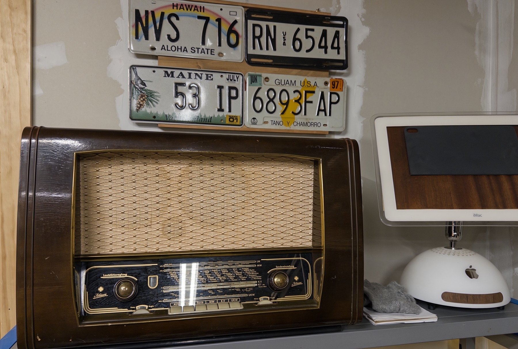 an old German tube radio on a shelf next to an old iMac with wood panel instead of a screen; above is a wall hanging with four license plates from places we have lived
