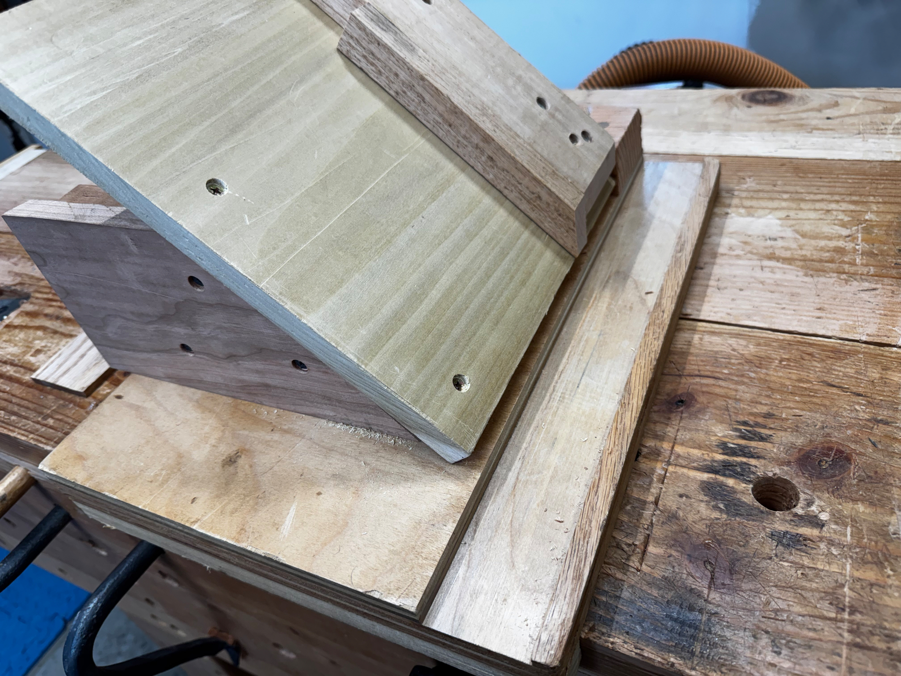 A wooden jig with angled pieces to shoot 45 degree miters is mounted on a workbench.