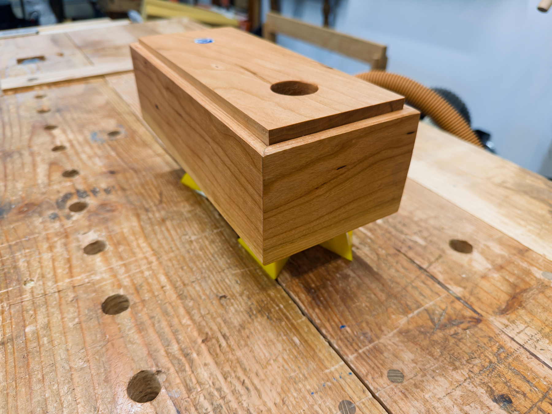 A wooden box with a circular hole on top rests on a workbench, supported by yellow stands.