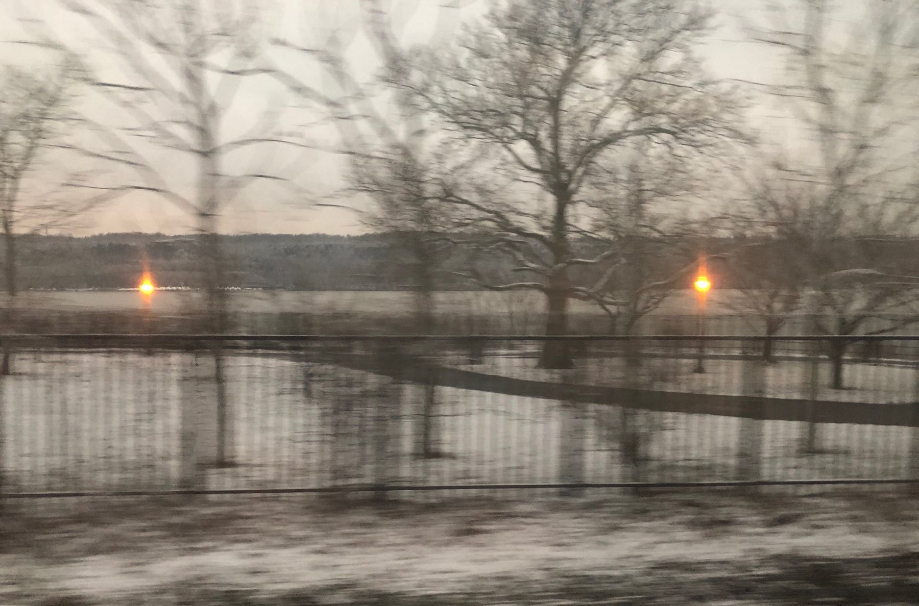 Snowy view of the Hudson river in the early morning from the train. Old fashioned streetlamps are still lit and the trees are bare.