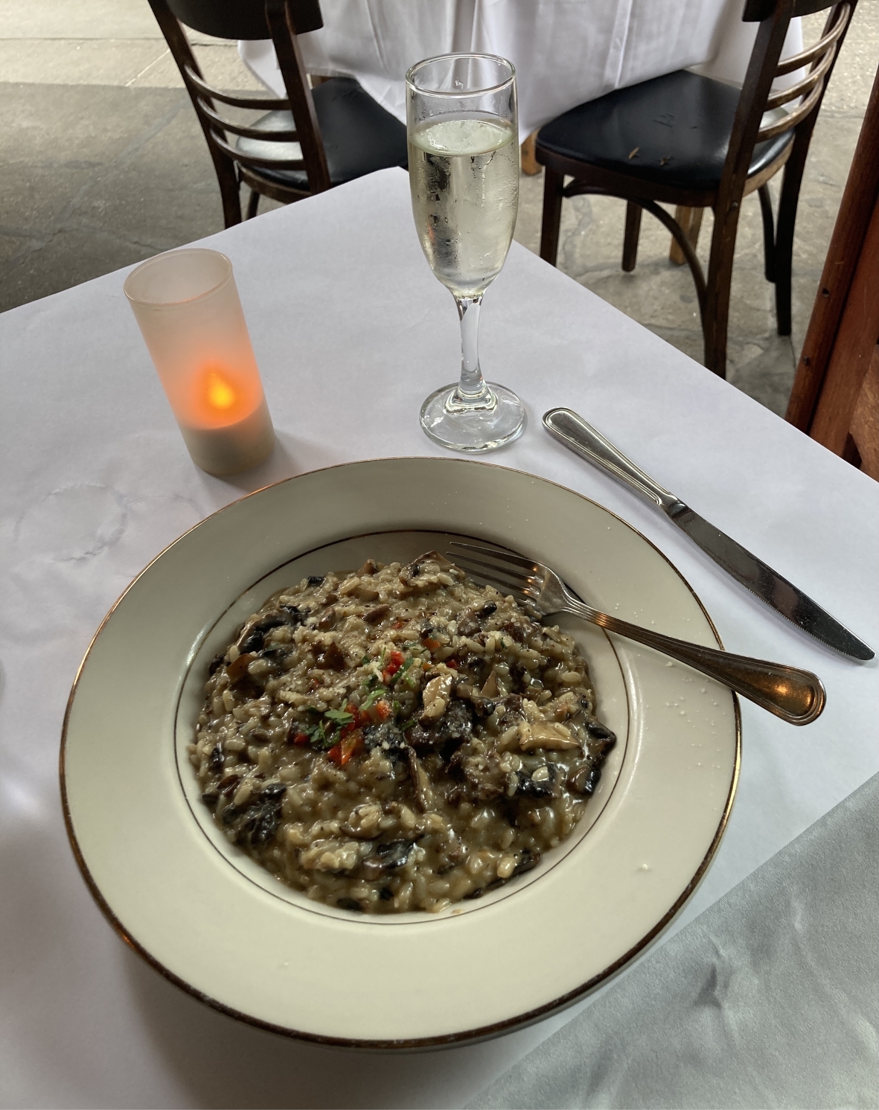 Large plate of mushroom risotto and a glass of prosecco on a white tablecloth. There's a fake LED candle and soft daylight is illuminating the scene. 