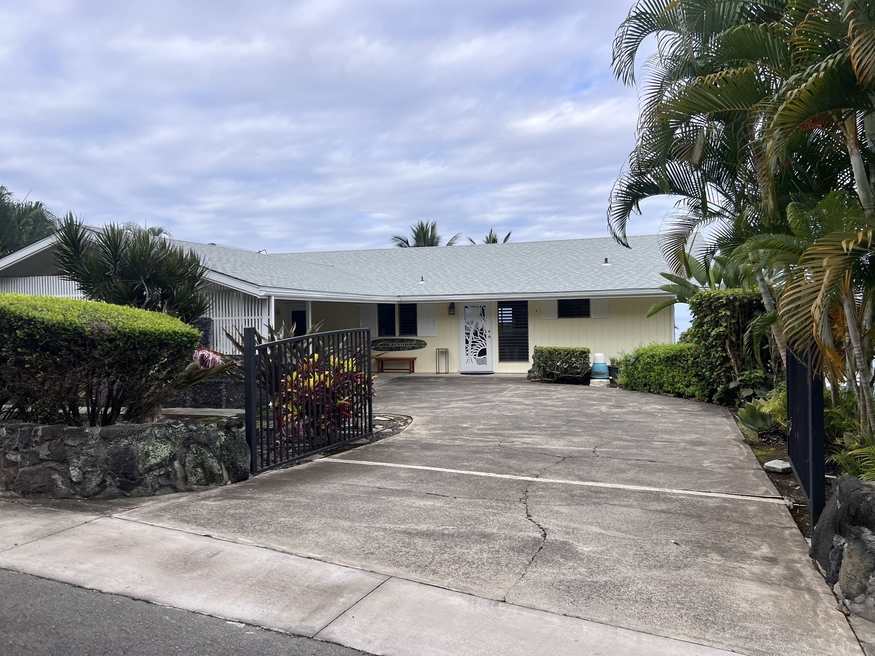 A single-story house with a light gray roof and a driveway, surrounded by tropical plants and a gated entrance, is set against a partly cloudy sky.