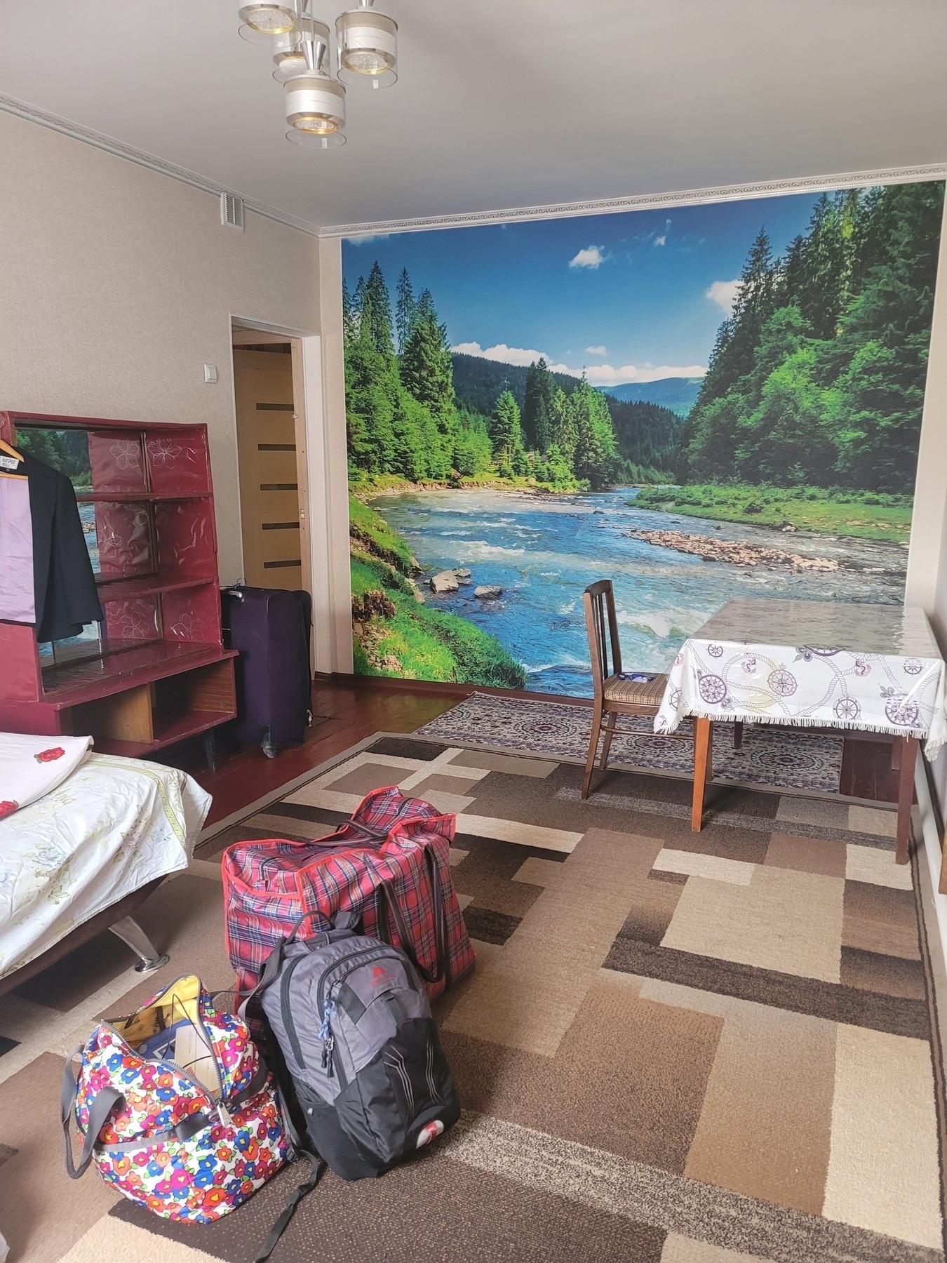 large bedroom with a river in forest wall mural on the back wall. shelf and bed on the left side of the room and a table and chair on the right side