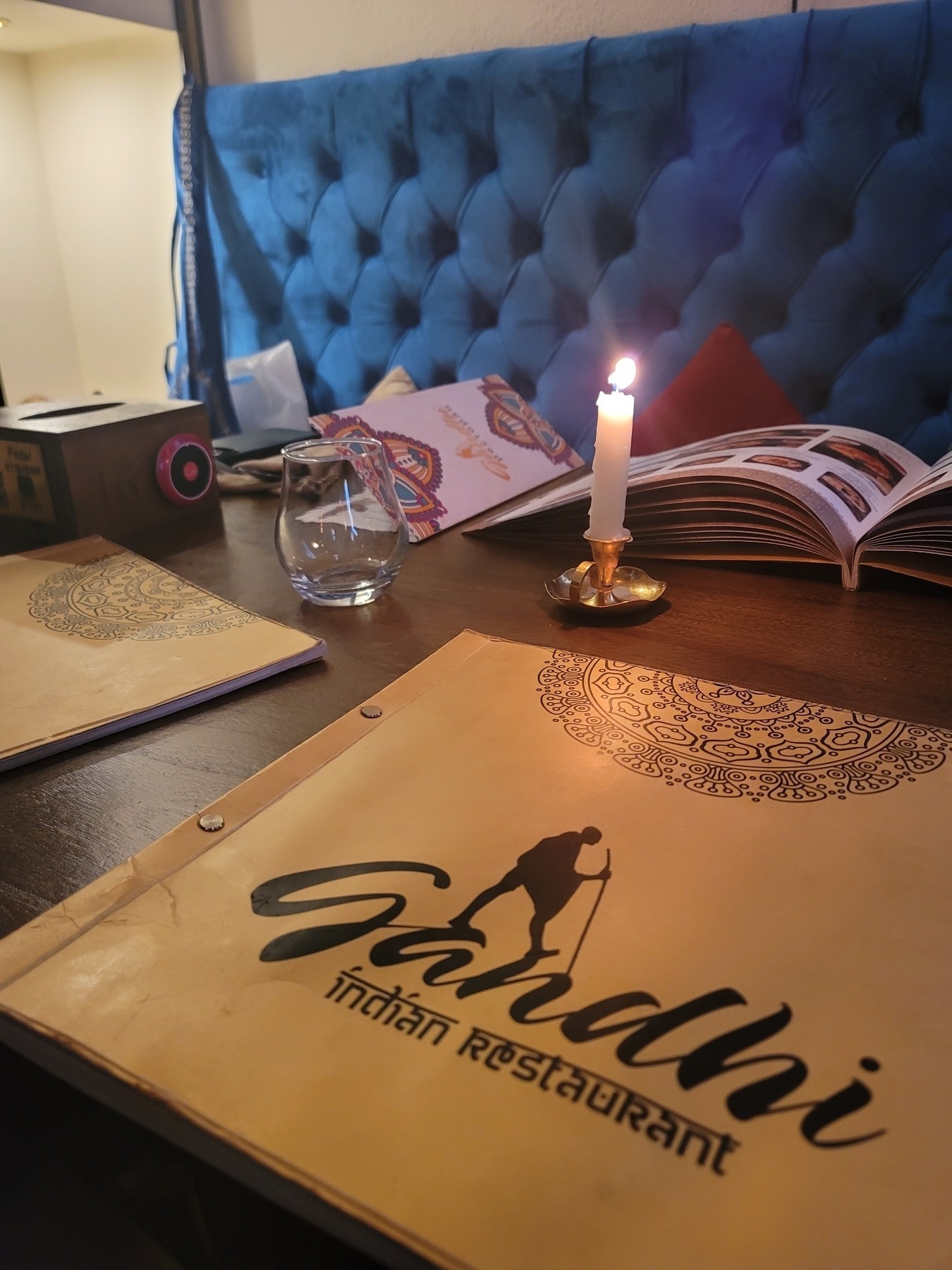table for four with a blue cushioned booth seat, 4 menus on the table with 2 open, 1 closed menu showing the name of the restaurant (Gandhi) and a candle in the center
