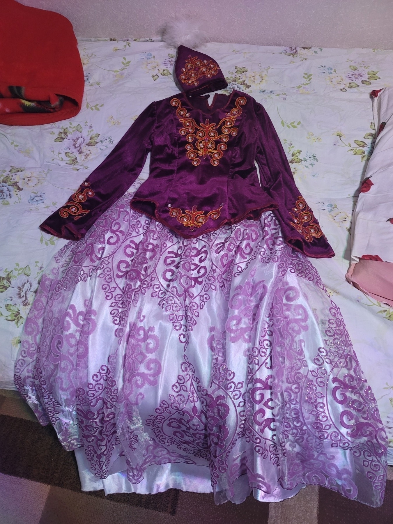 traditional female Kyrgyz clothes - long skirt with white silk slip and a mesh outer layer with a purple pattern; purple velvet-like top with red/orange embellishments; matching purple hat with red/orange embellishments and white feathers from the top