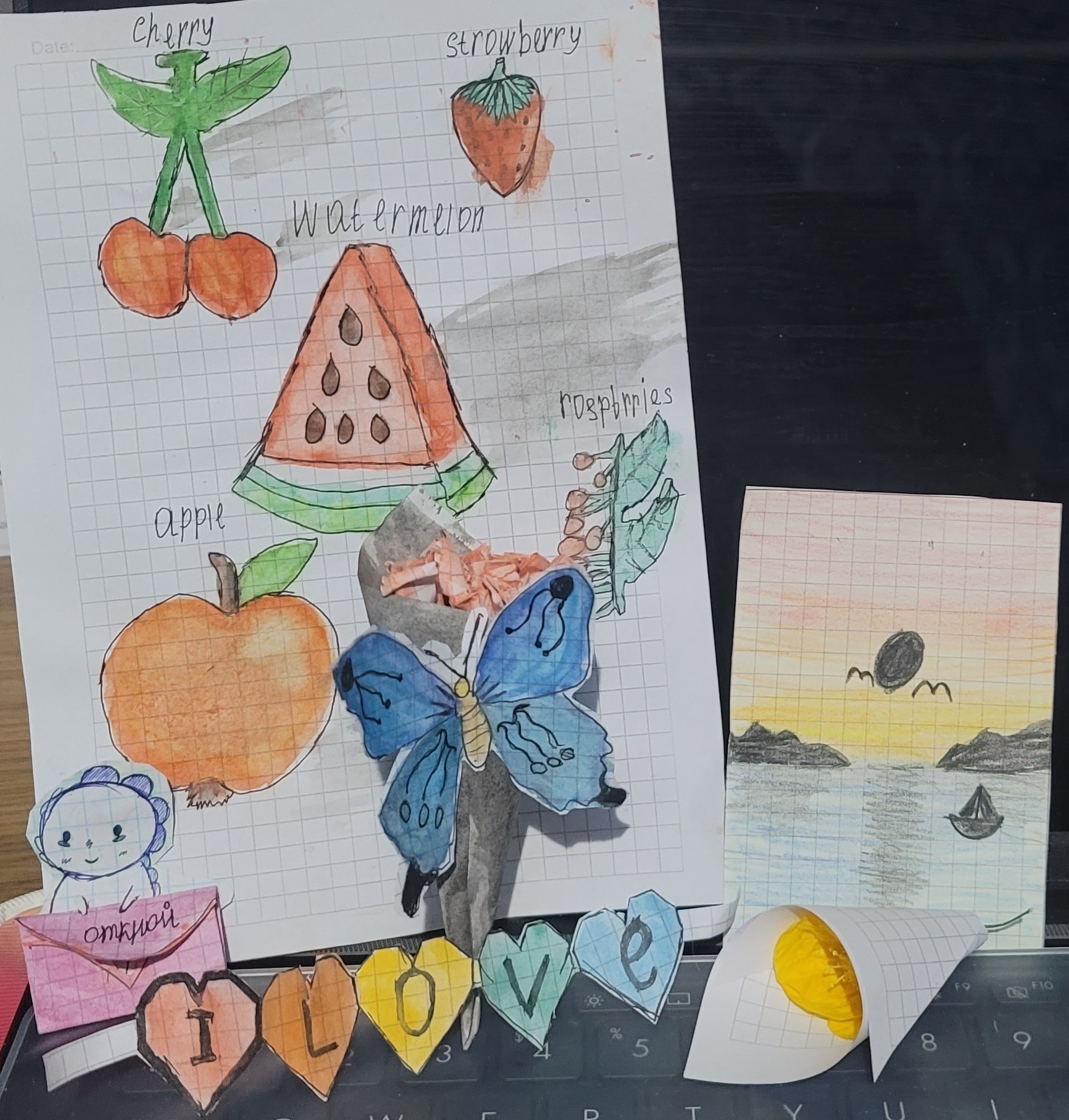 one paper with colored pictures of different fruits labeled in English; picture of a boat on an ocean at sunset; paper bouquet of paper roses with a blue paper butterfly on it; real, small yellow flower in a paper bouquet; I L O V E, each letter in a different colored heart; a drawn dinosaur holding a pink paper envelope which reads 'open' in Russian