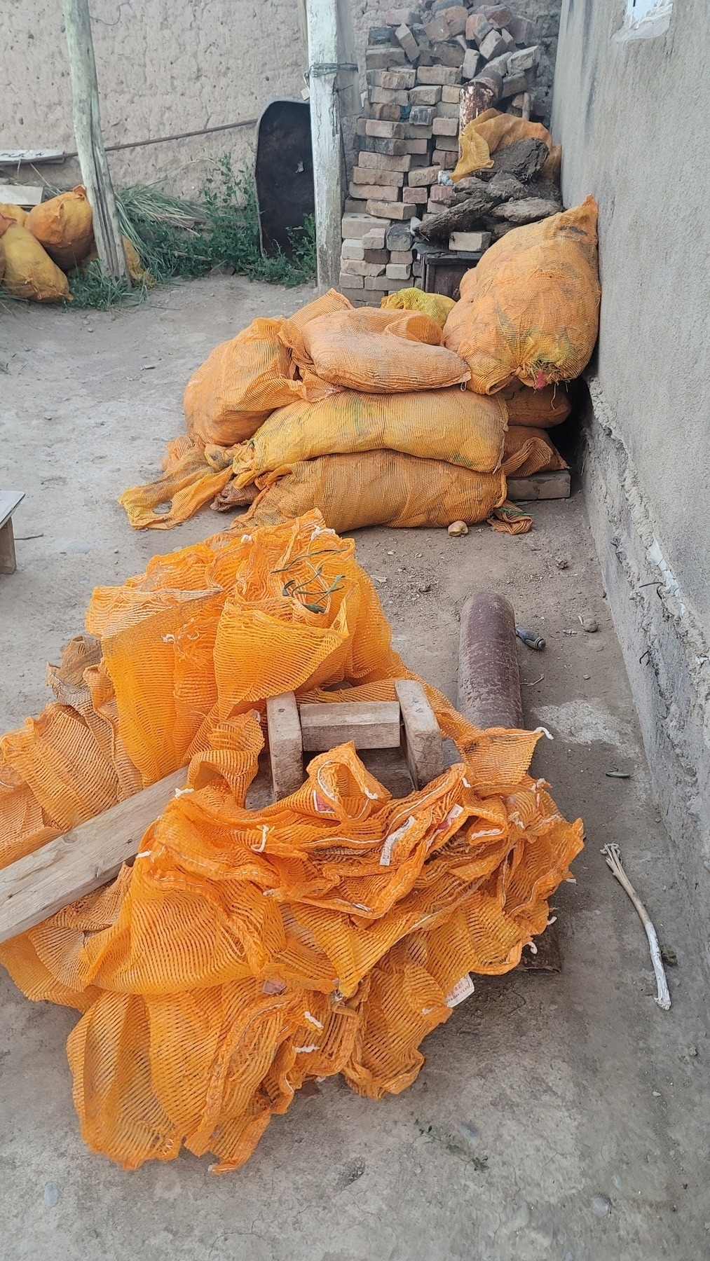 unfolded orange mesh bags in a pile under a plank of wood since it's windy here; in the background, bags filled with more bags that have yet to be unpacked