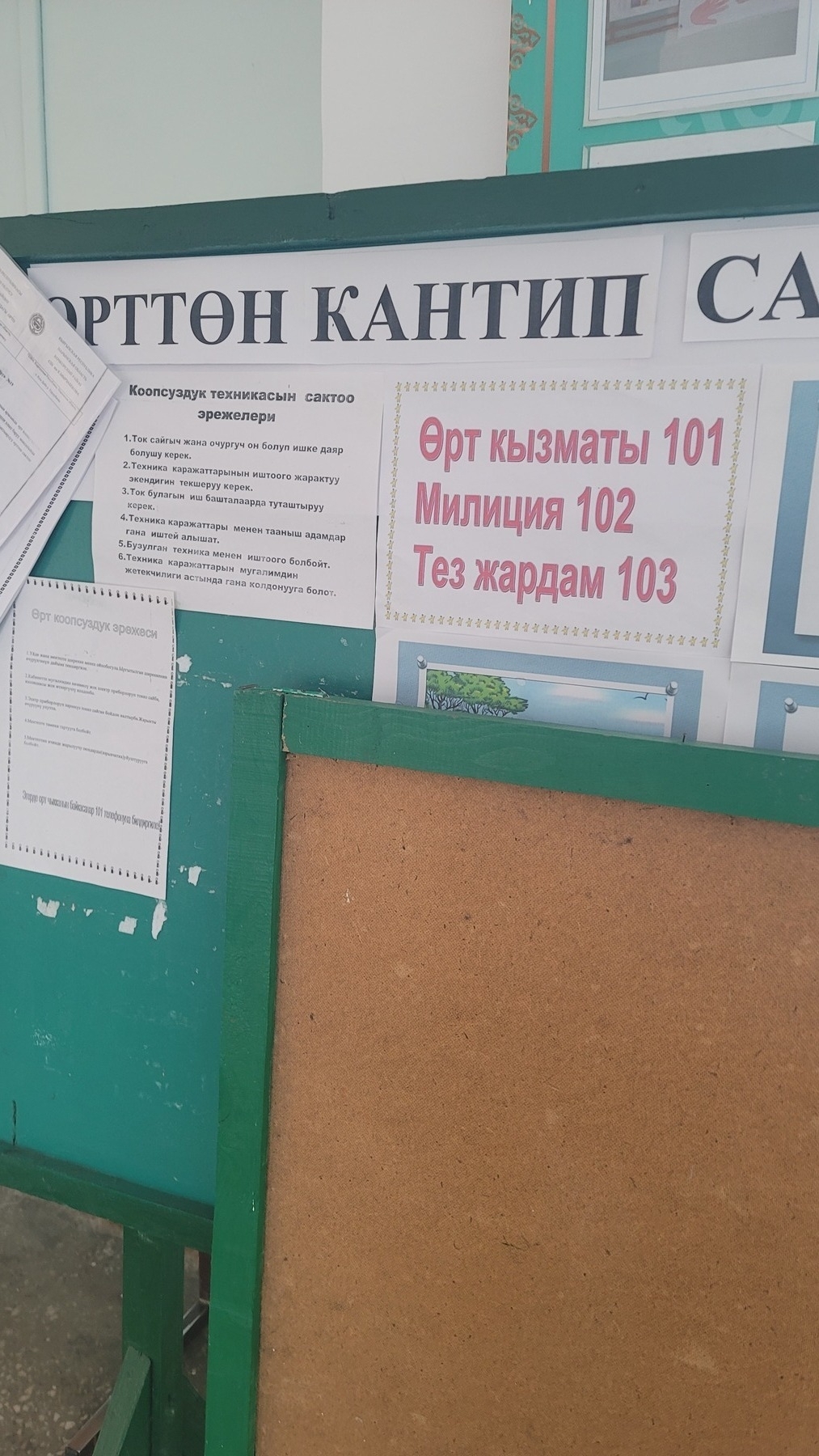 green bulletin board with several white papers with Kyrgyz text taped to it, including one with emergency number descriptions in red text