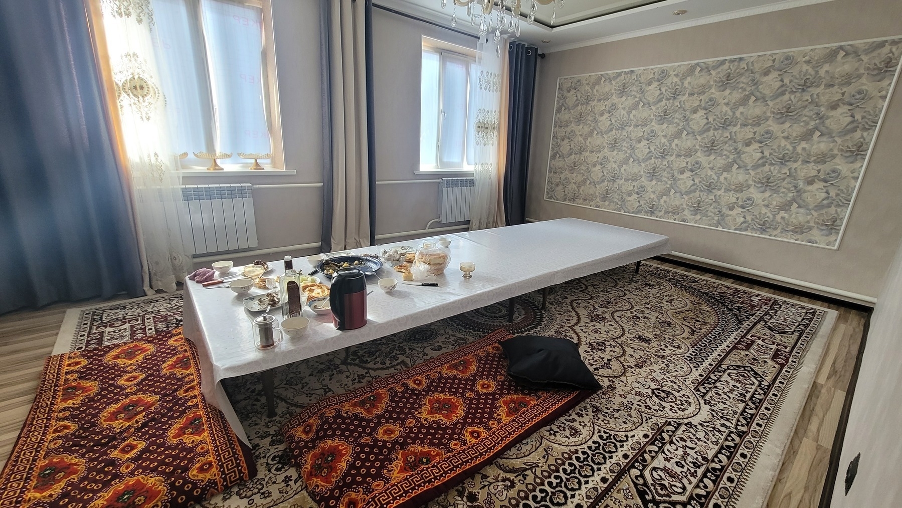 big room with a long table (white table cloth on top) with two traditional Kyrgyz rugs spread along one short side and one long side as seating. bread, honey, and tea among other things on the table