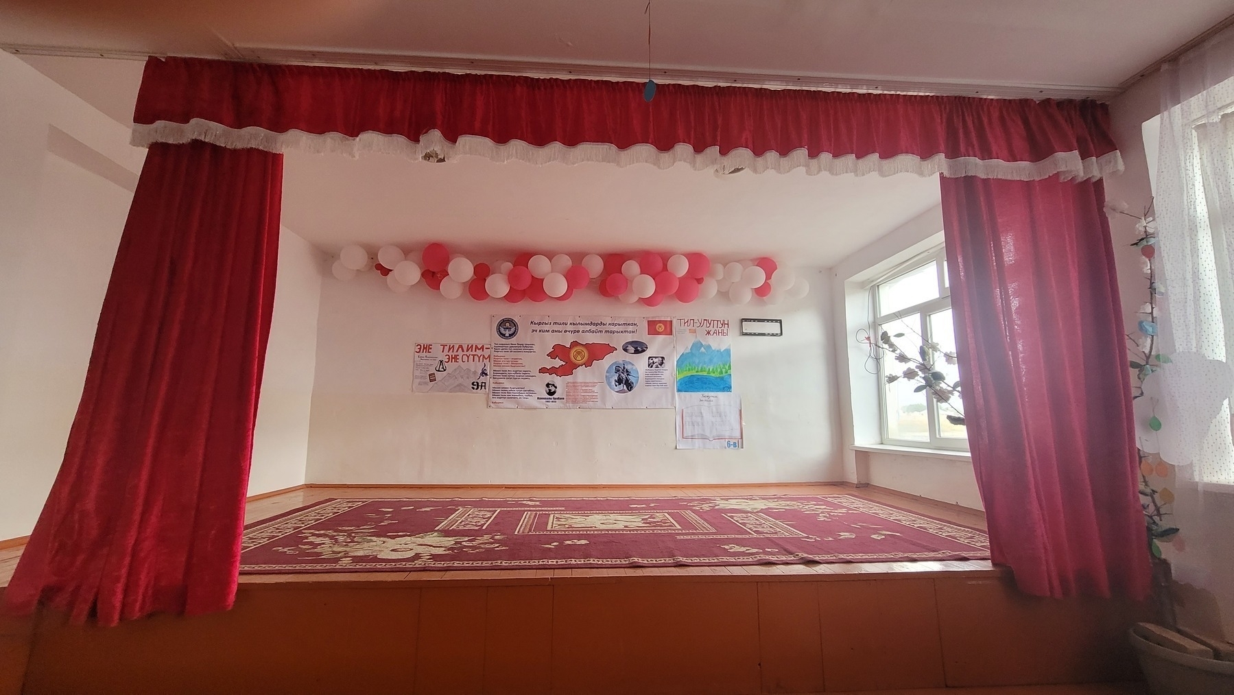 stage where students performed. brown/orange platform with a red rug on it, curtains framing it; red, pink and white balloons on the wall at the back of the performing area and hand-drawn student posters about Kyrgyzstan beneath those