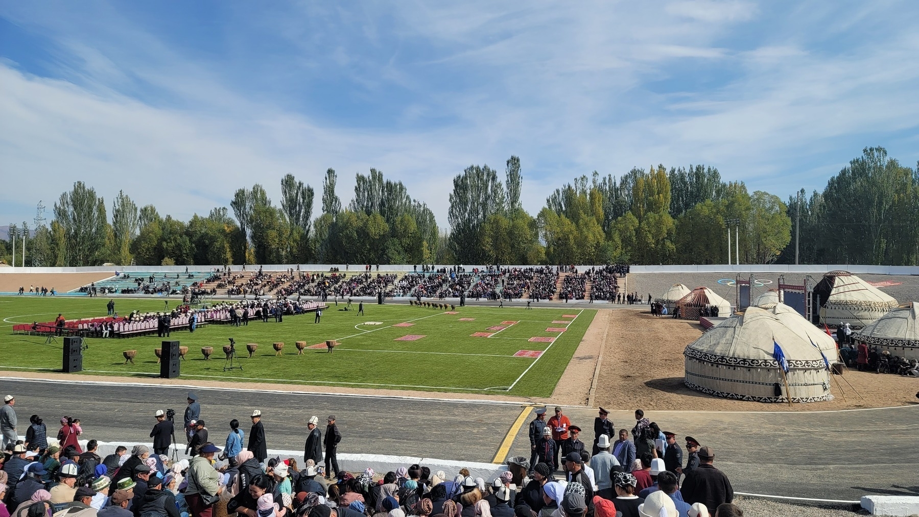 looking down onto a football field in a stadium with spectators sitting on bleacher benches and in chairs on the field pointed toward one of the ends of the field, where about 7 yurts are set up