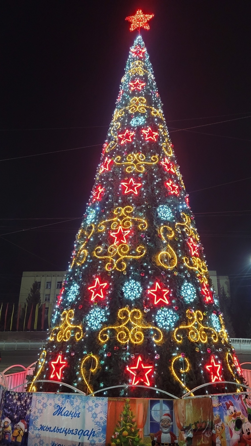 tall Christmas tree with decorative light patterns and ornaments. star at the top. sign in front which reads "Happy New Year" in Kyrgyz