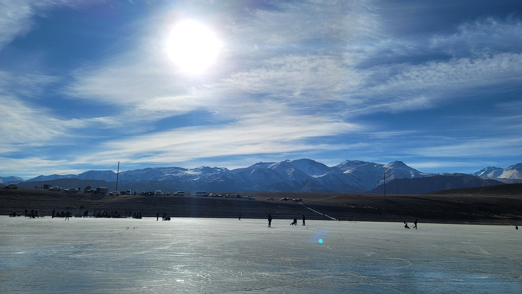 frozen lake with some ice skaters, a brown hill above with parked cars, and snow-capped mountains in the distance