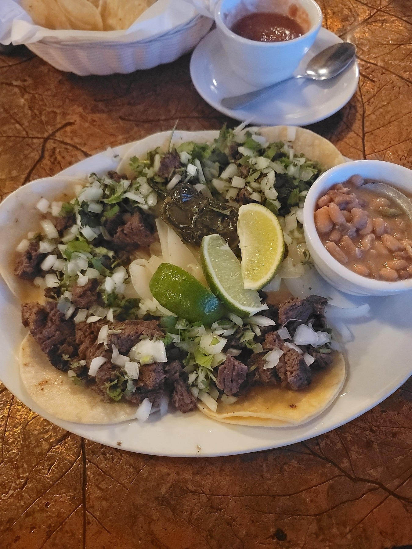 plate of 5 small beef street tacos topped with onion and cilantro, a green pepper and a few lime wedges in the middle, and a small side dish of beans on the right