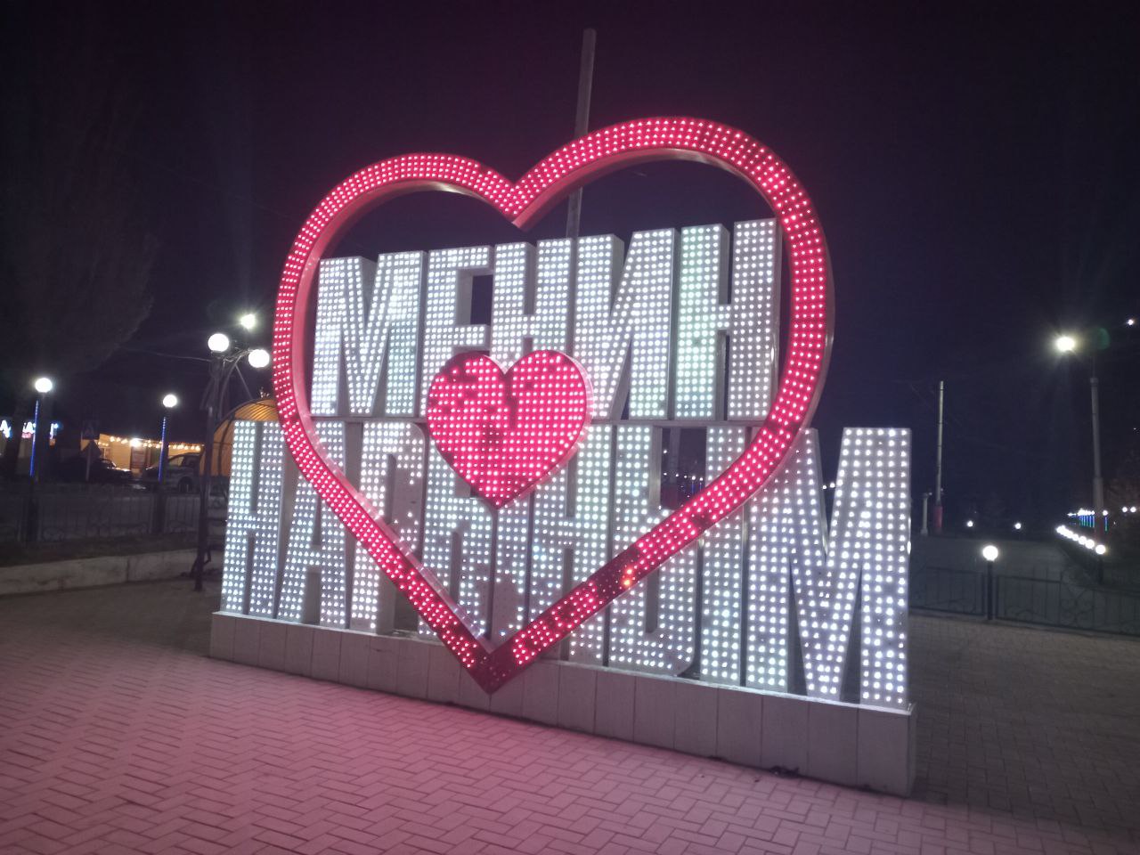 a standing, illuminated sign with a red heart surrounding white text which says, "My Naryn" in Kyrgyz in a small city in the evening