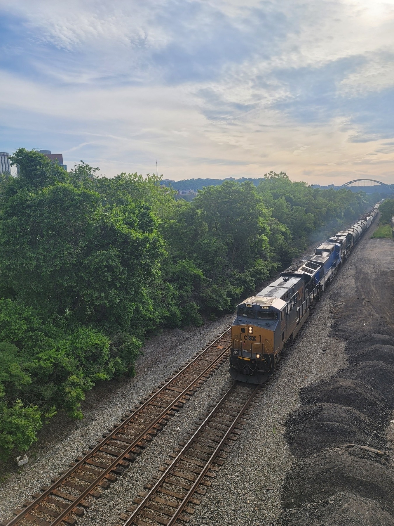 incoming train on tracks seen from above headed towards bottom left of image; green trees on the left and piles of dirt on the right