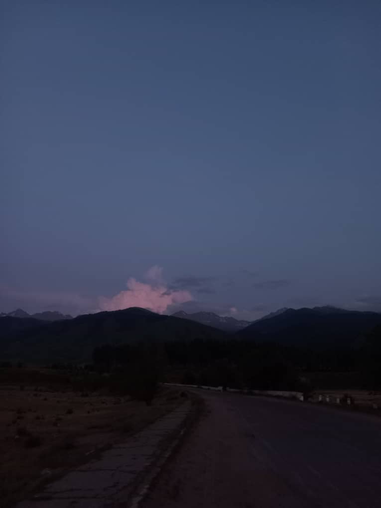 gravel road curving slightly towards green mountains in distance with a light pink cloud above at dusk