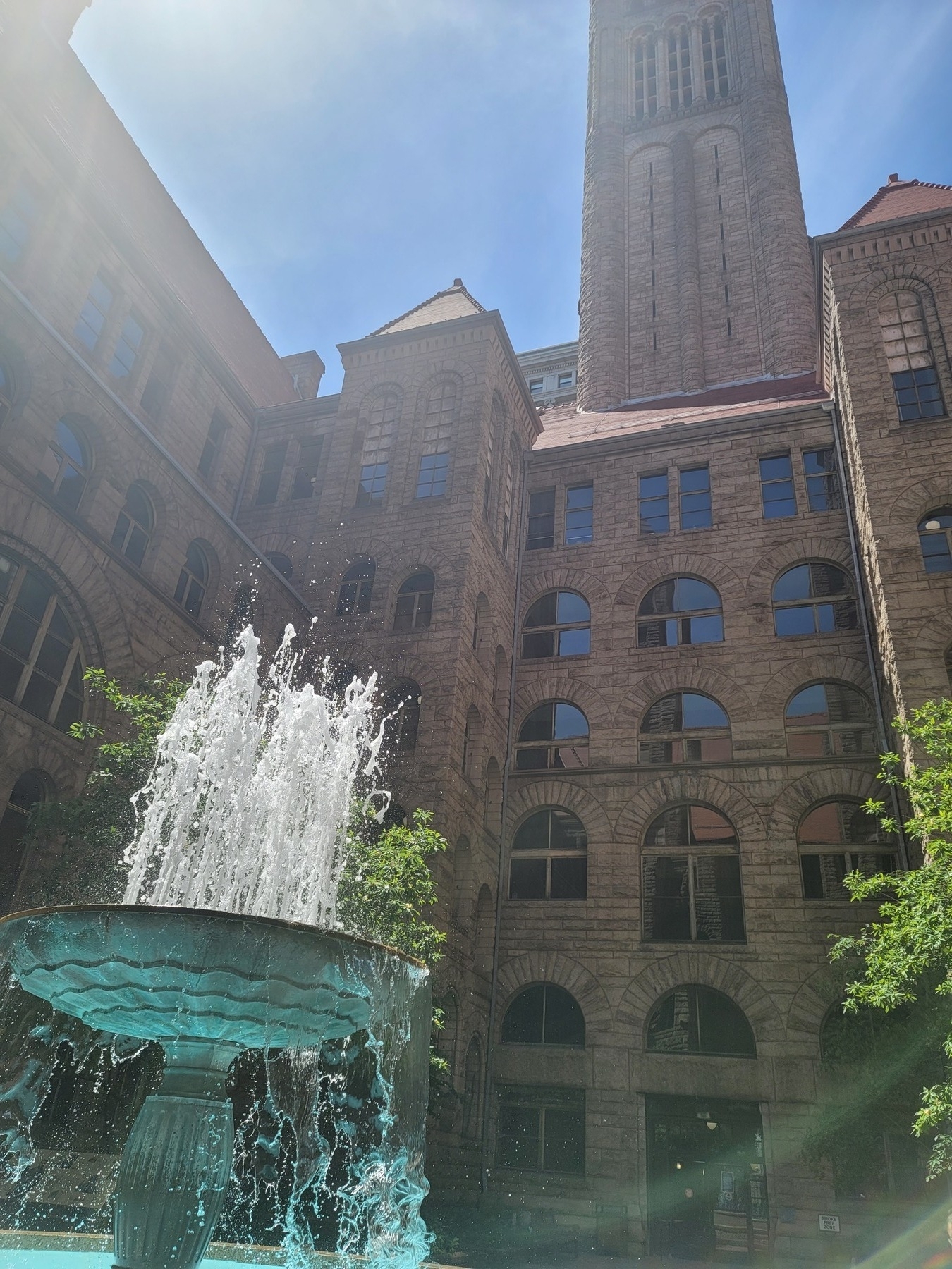 fountain in lower left corner with water spurting from the top in front of a brown, romanesque revival style building