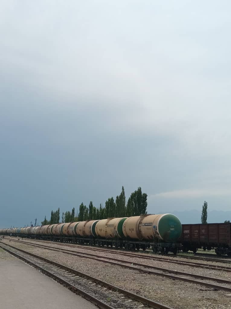 green and light yellow-ish petrol train(?) moving towards left side of photo with a brown train parked on a different rail behind it