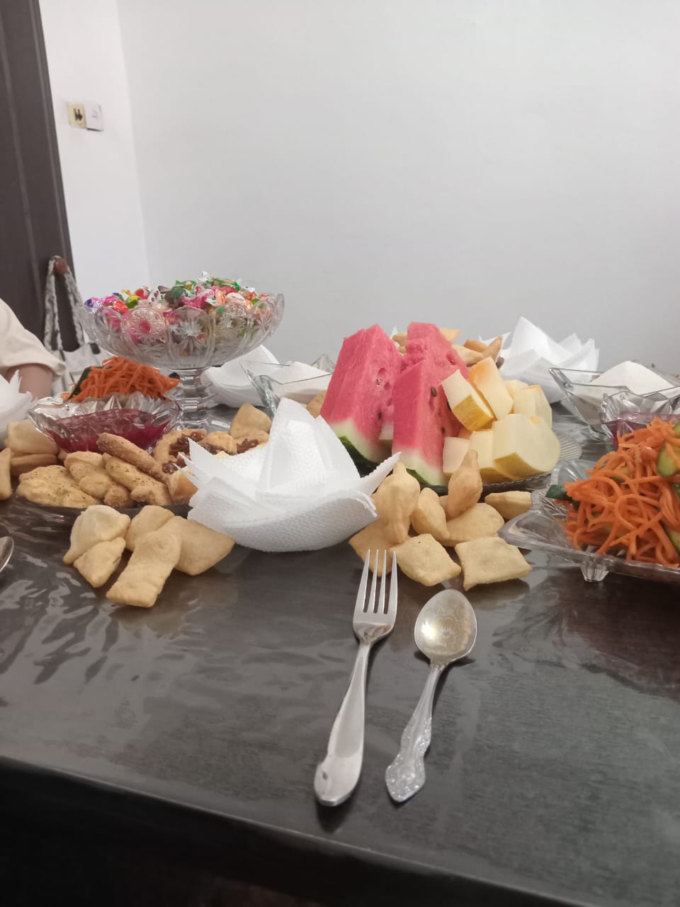 tabletop set with cutlery, napkins, sliced watermelon, melon, carrot and cucumber salad, candy, cookies and small pieces of fried bread