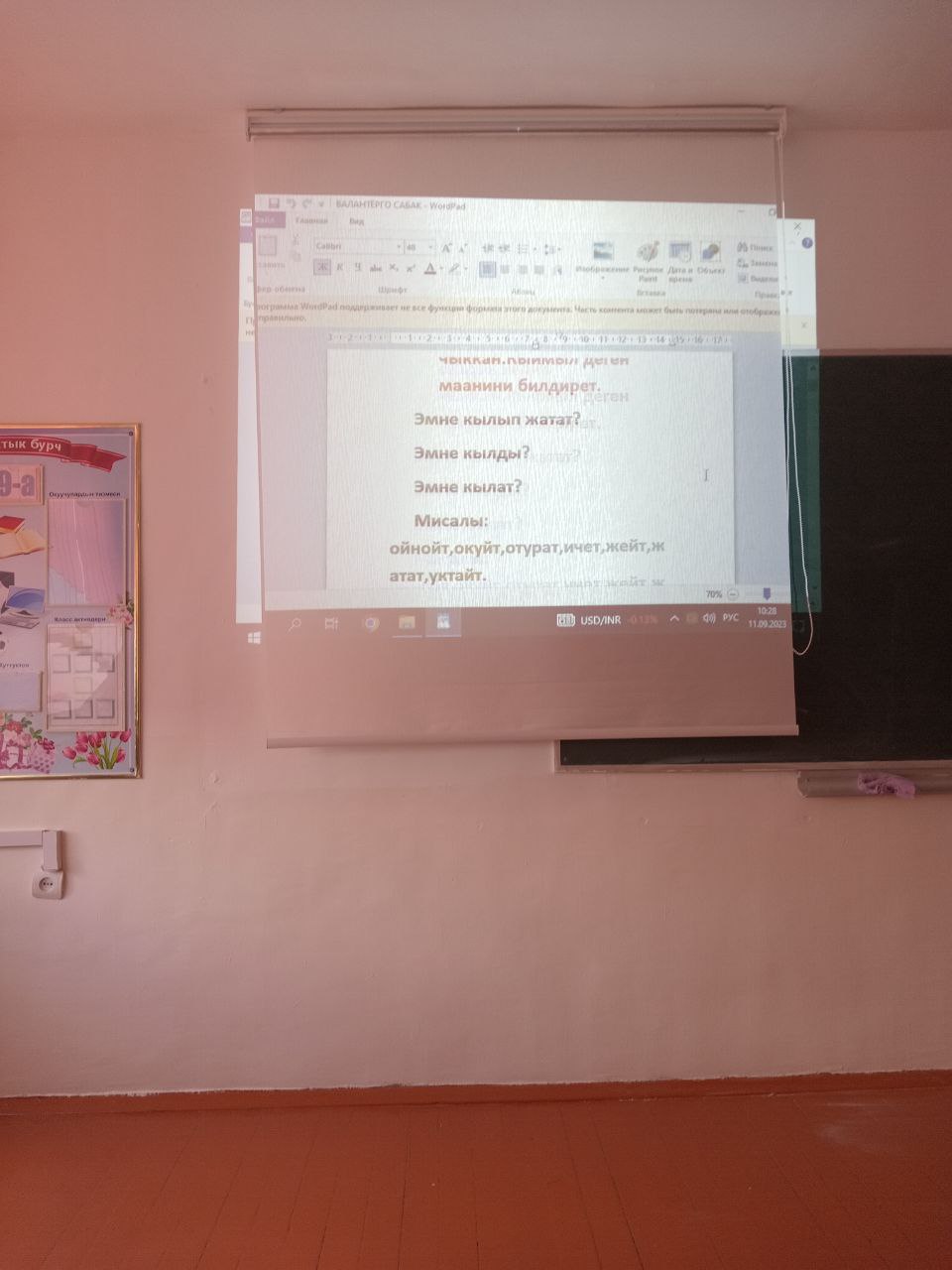 A Microsoft Word document being projected onto a roll-down overhead screen in a classroom. The Word doc has Kyrgyz words on it