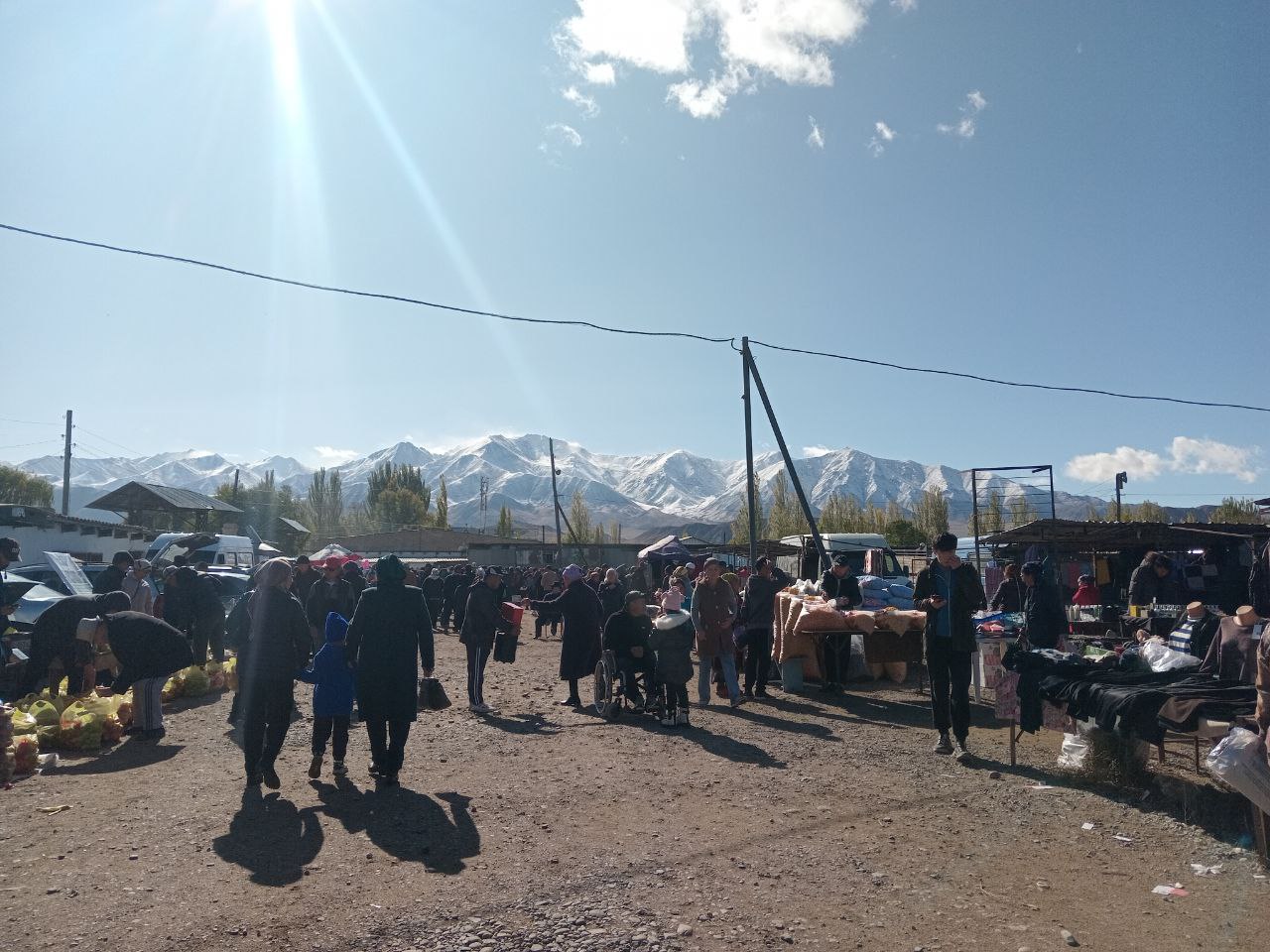 people walking around, tables and booths set up along a dirt road with people selling various things including food, power lines and mountains in the background